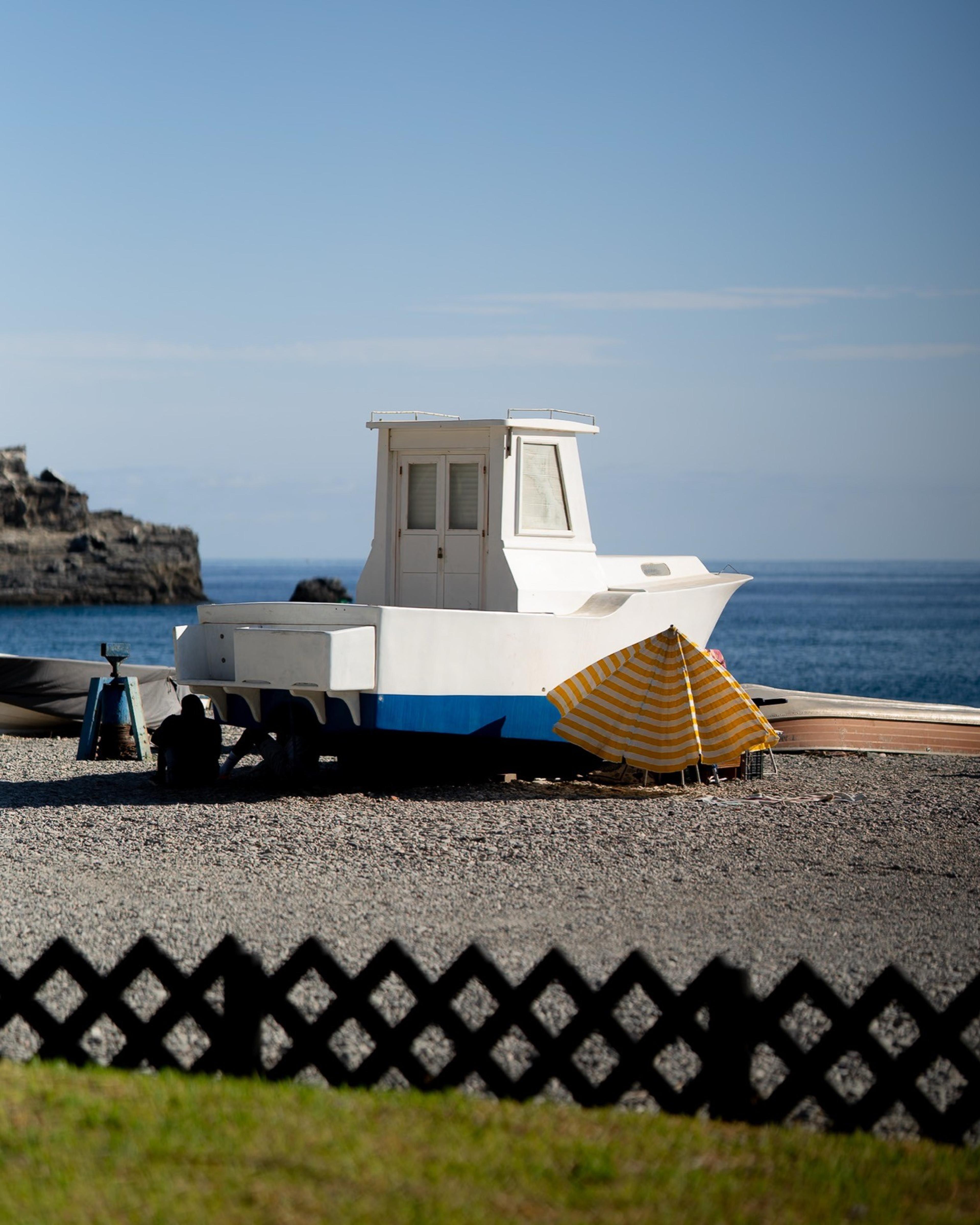 White fishing boat resting on a pebble beach beside a yellow striped parasol by the sea