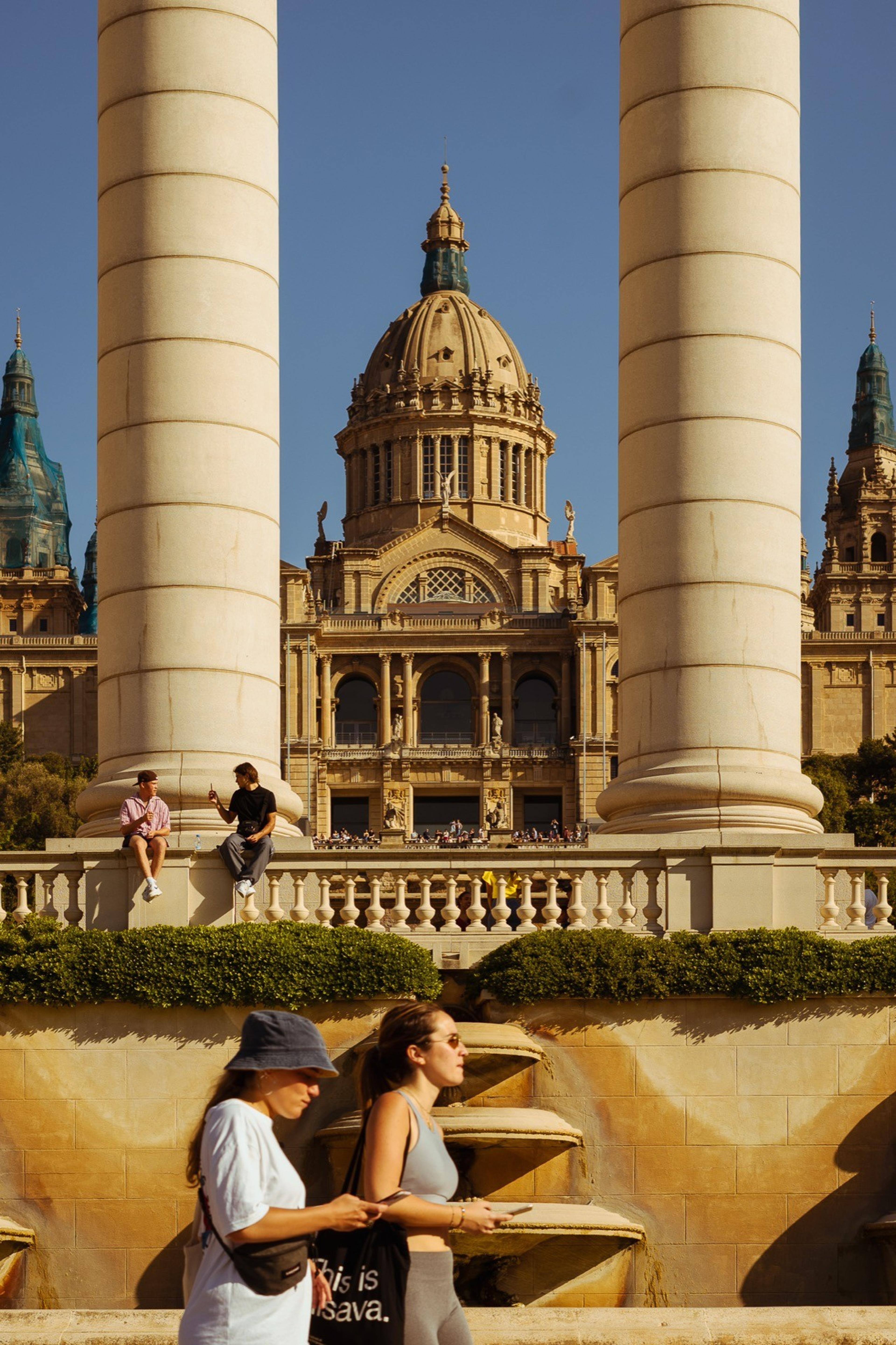 Two women walking past a fountain between tall columns in front of an ornate domed palace