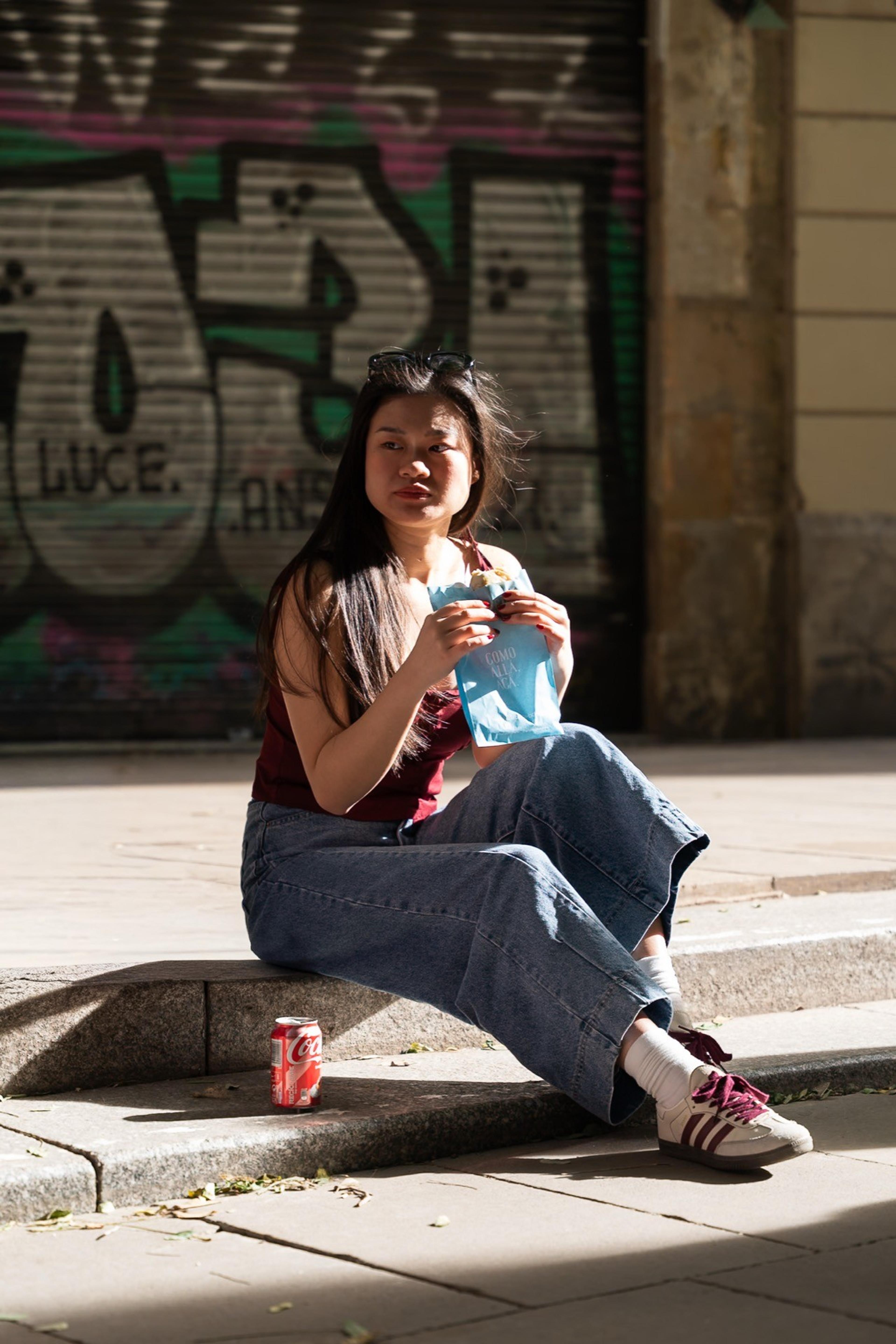 Woman in wide jeans sitting on a sunny curb eating, a graffiti-covered shutter behind her