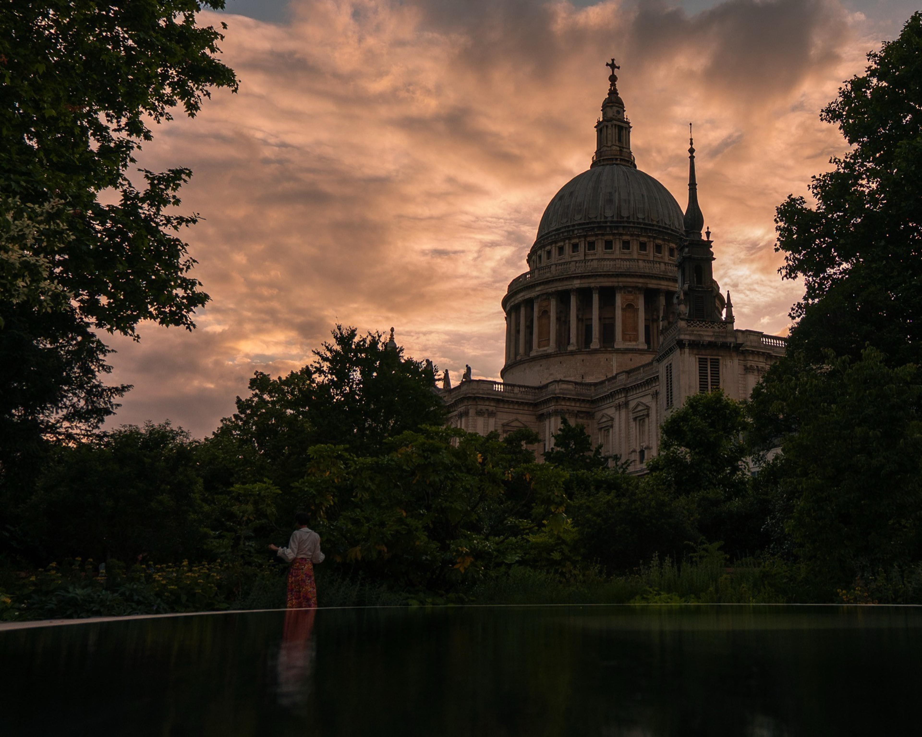 Domed cathedral silhouetted against a pink sunset sky, a person reflected in a garden pool
