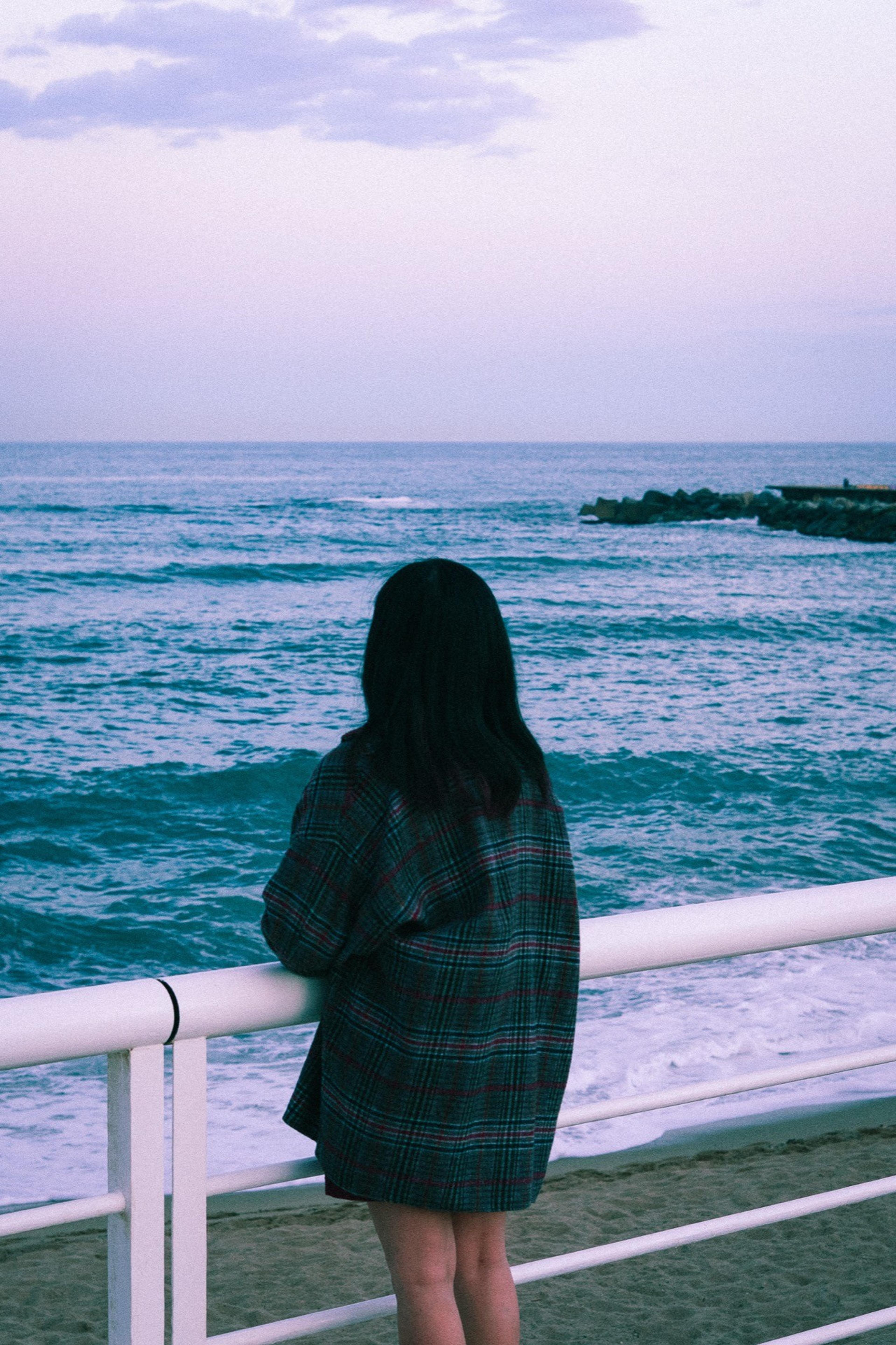 Woman in a plaid coat leaning on a white railing, looking out at a turquoise sea at dusk