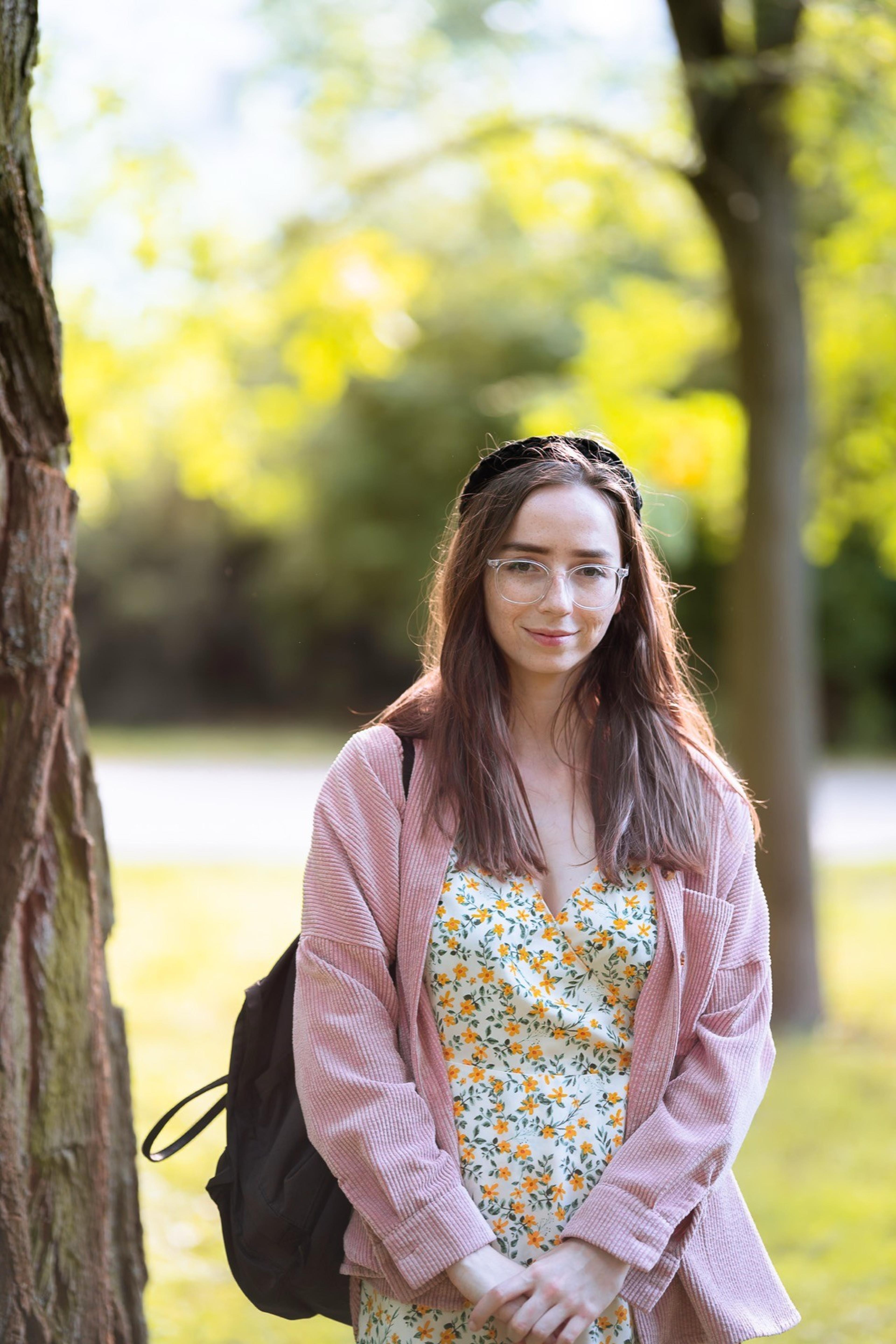 Smiling woman in glasses, pink jacket and floral dress standing beside a tree in a sunny park