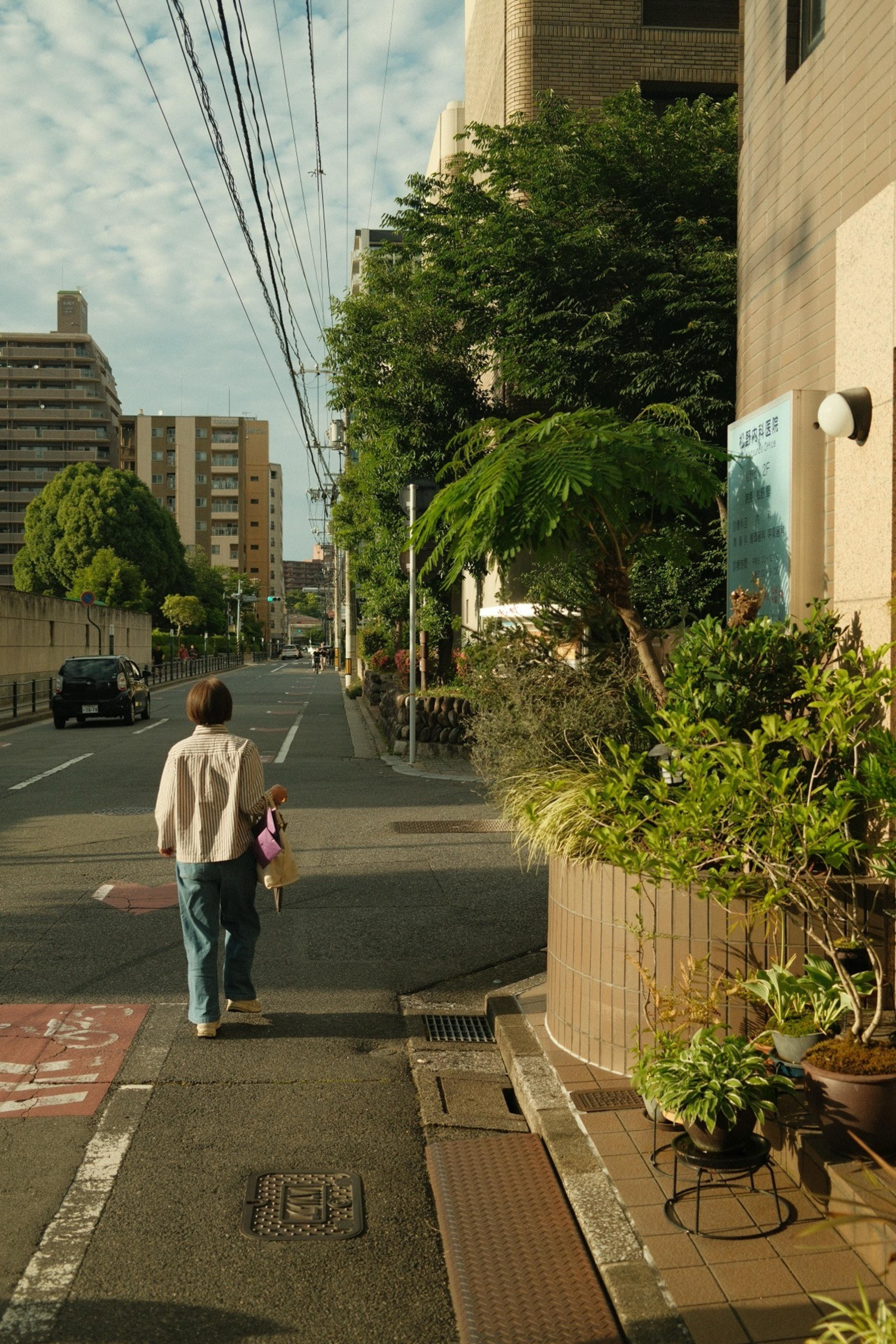 Woman walking along a quiet residential street lined with trees and potted plants in morning light