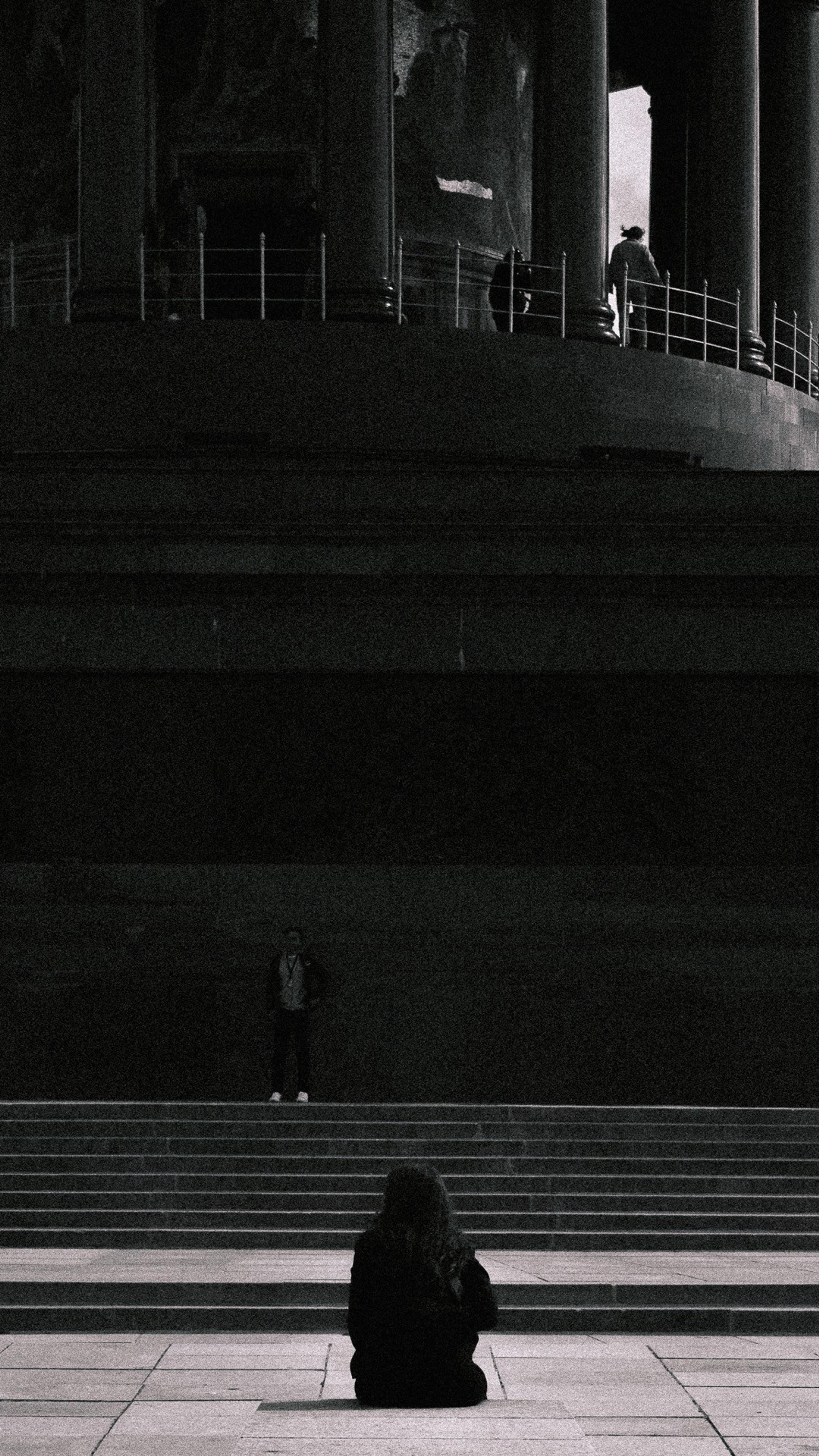 Black and white shot of a person sitting alone on wide stone steps below a colonnaded monument
