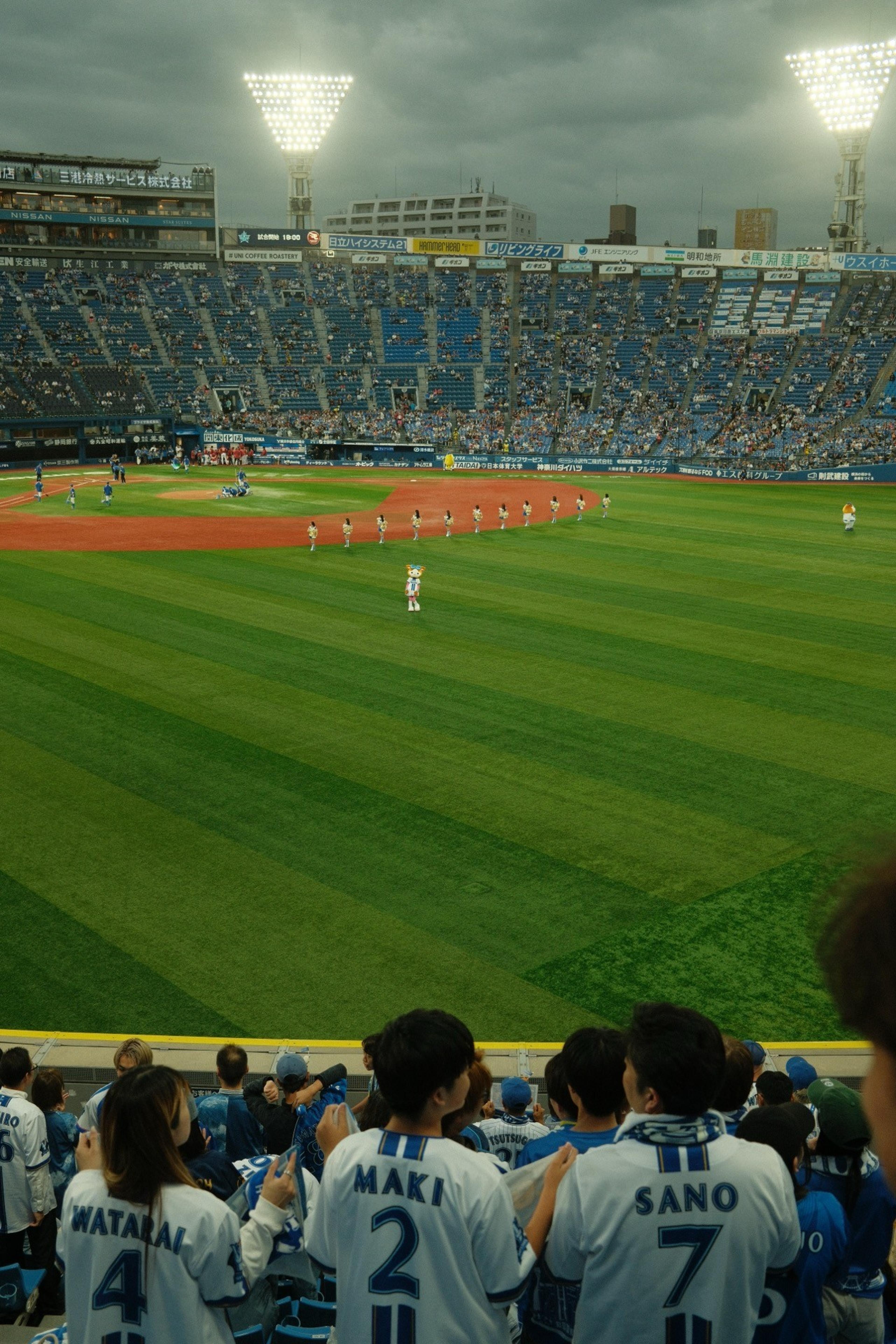 Baseball stadium at dusk with players lined up on the field and fans in numbered jerseys in front