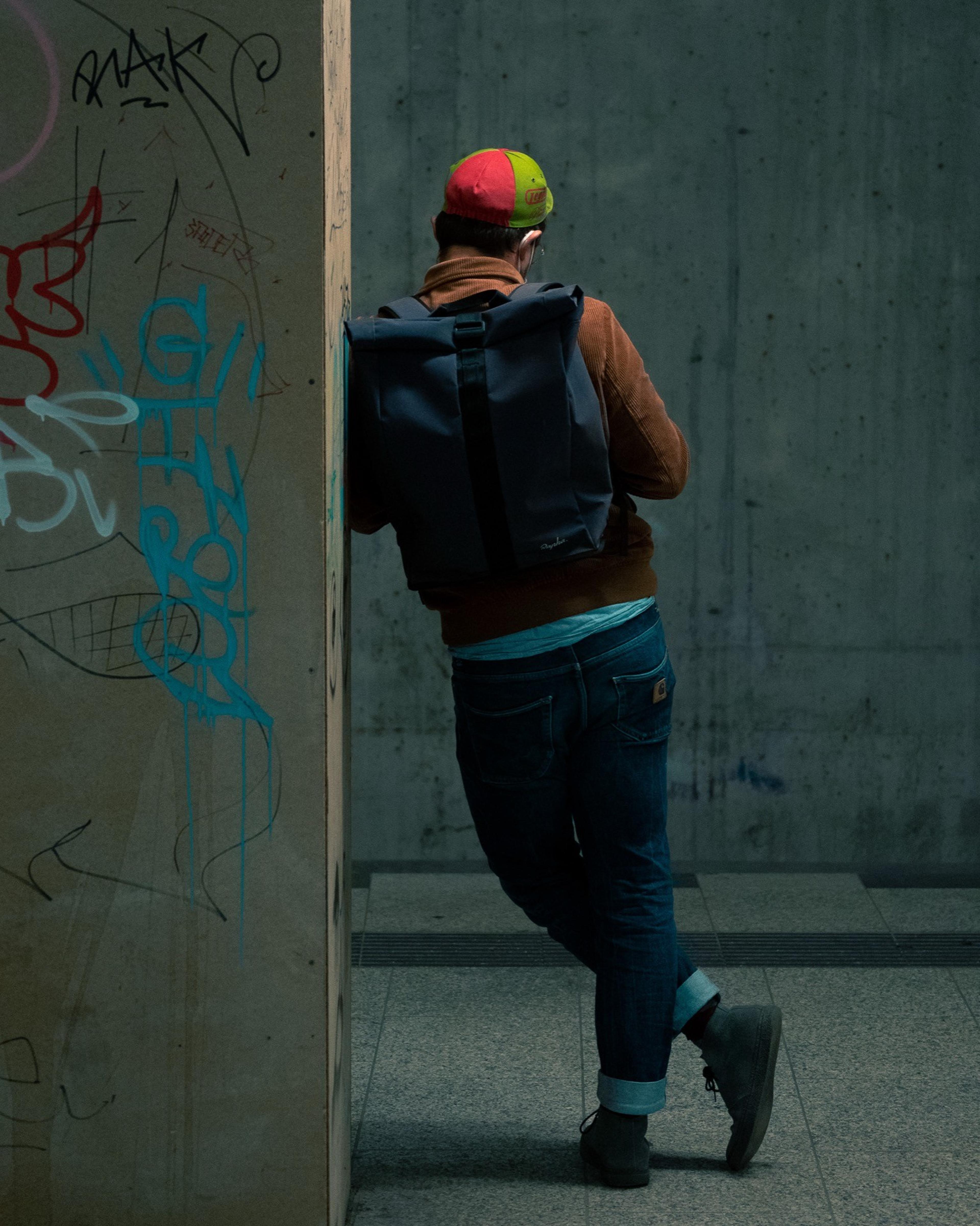Man in a colorful cap and backpack leaning on a graffiti-covered concrete pillar