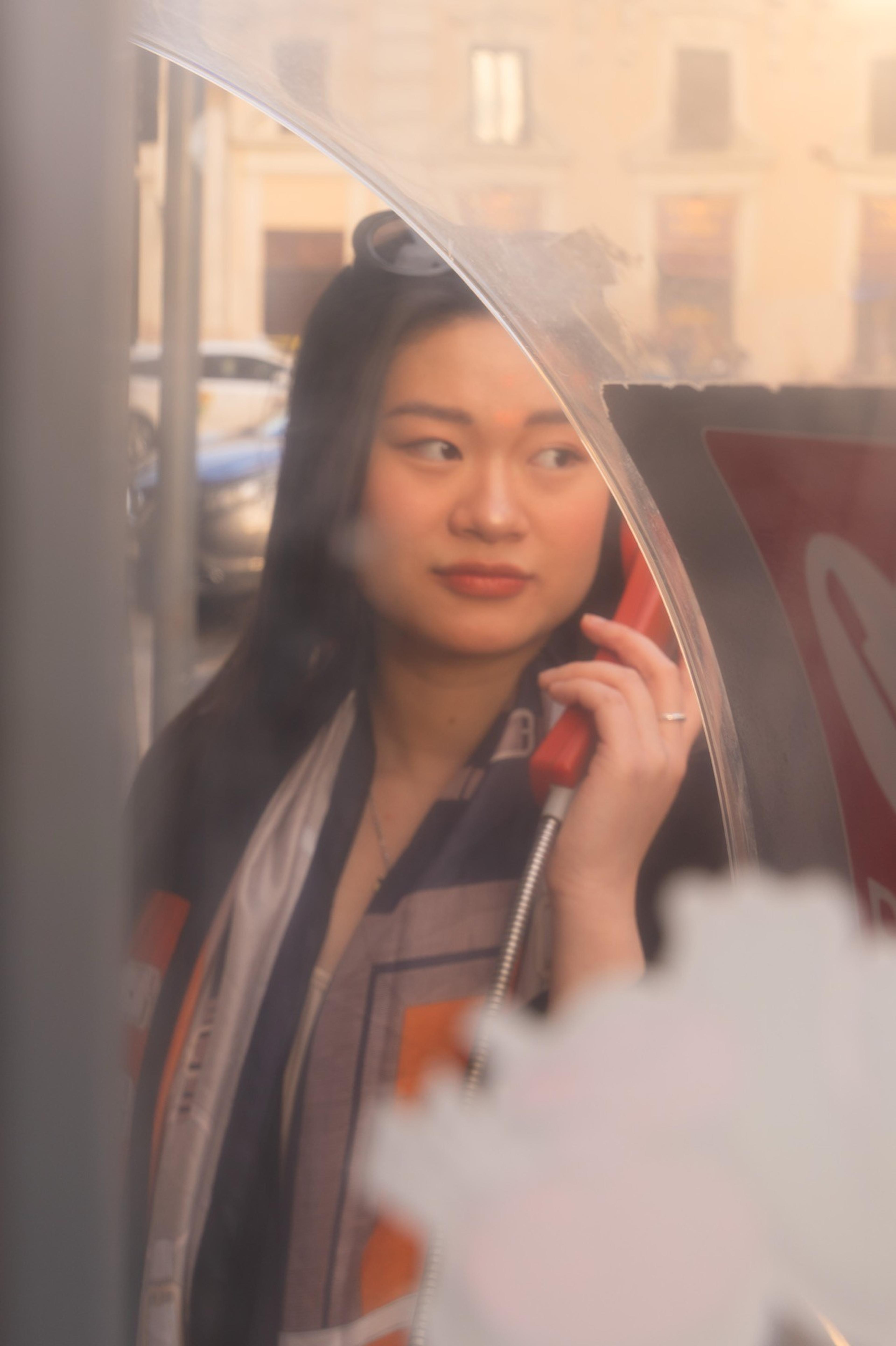 Woman holding a red handset inside a phone booth, seen through scratched glass in warm light