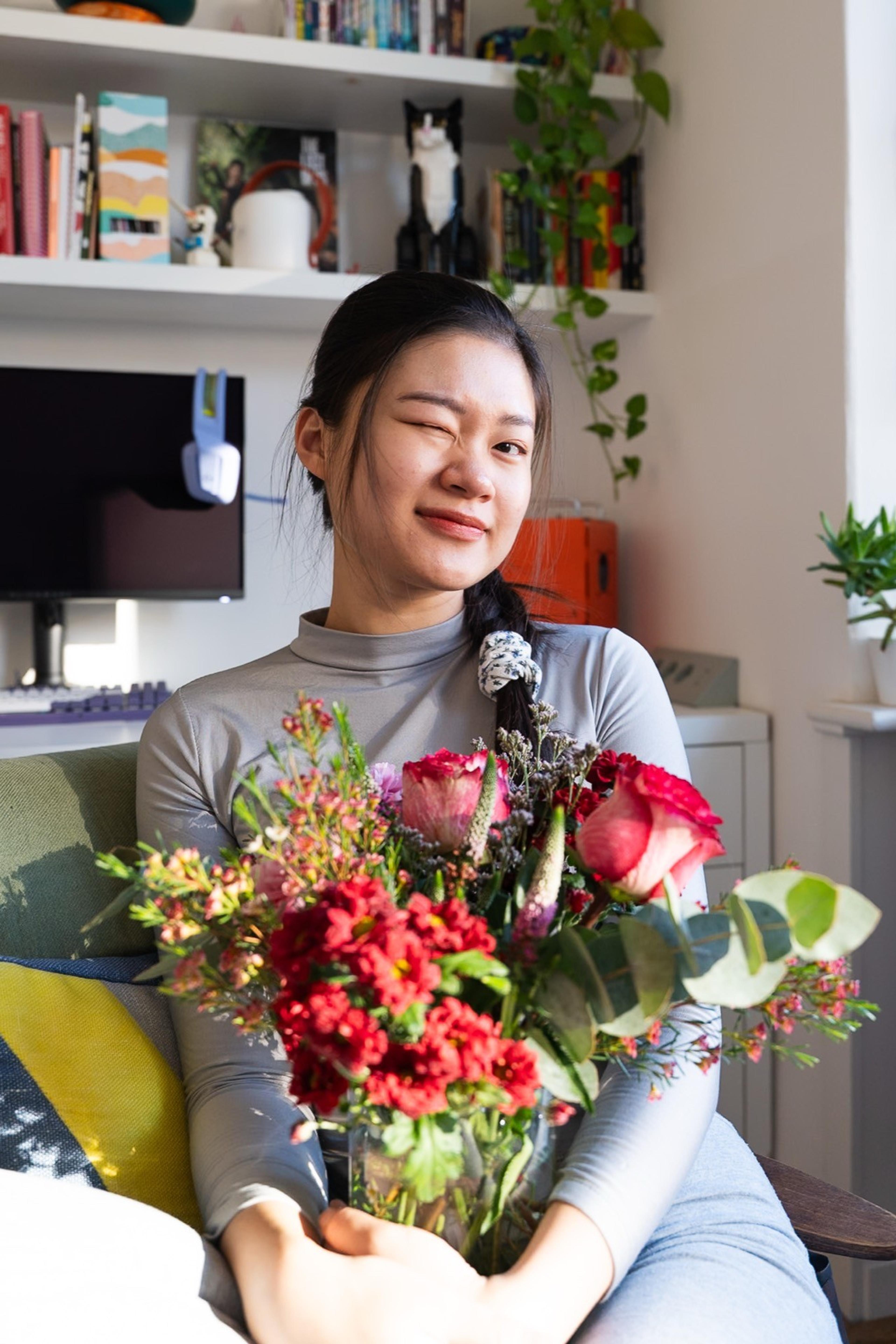 Woman winking while holding a vase of red roses and carnations at home in front of bookshelves