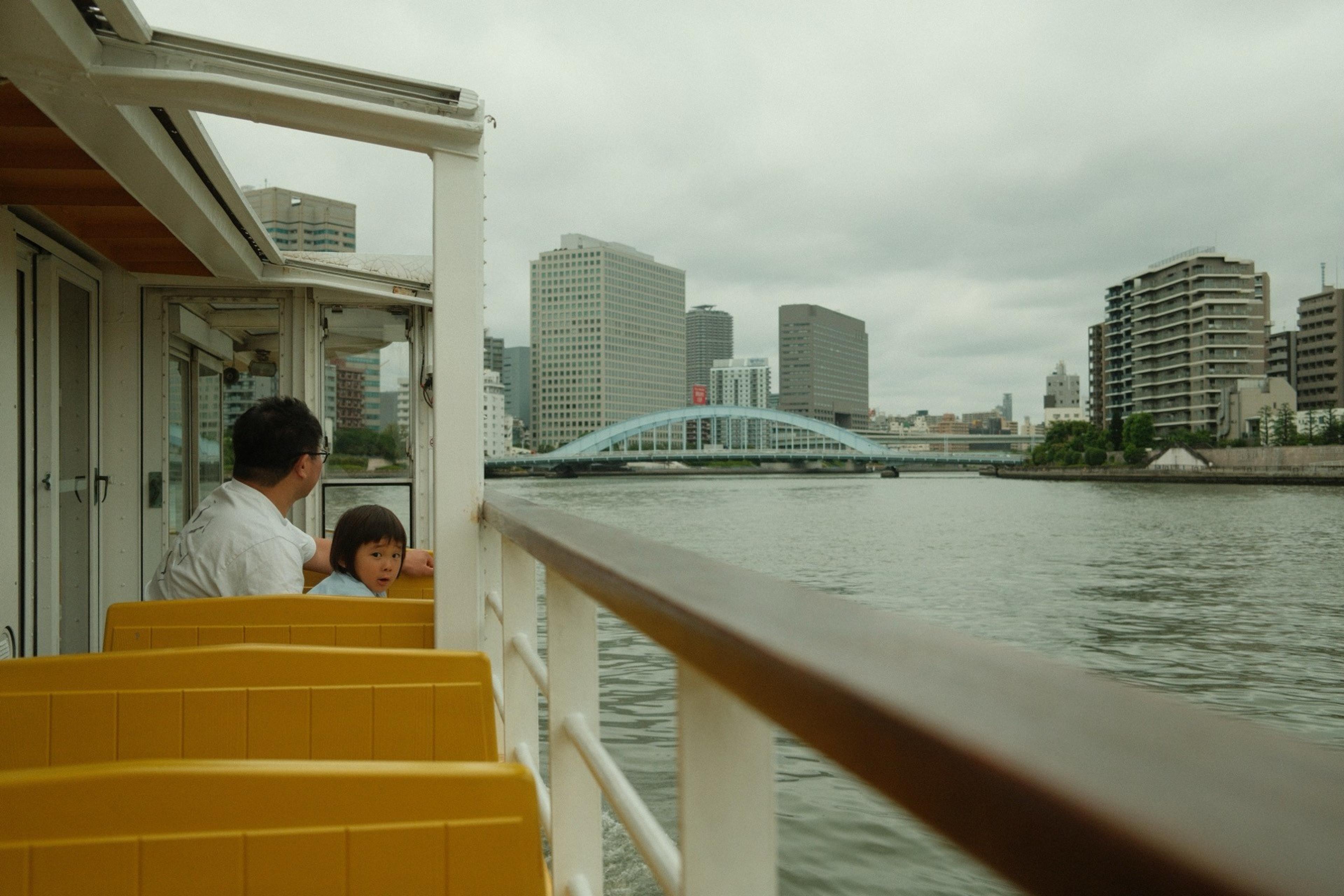 Father and child on a river boat deck with yellow seats, a blue arch bridge and high-rises ahead