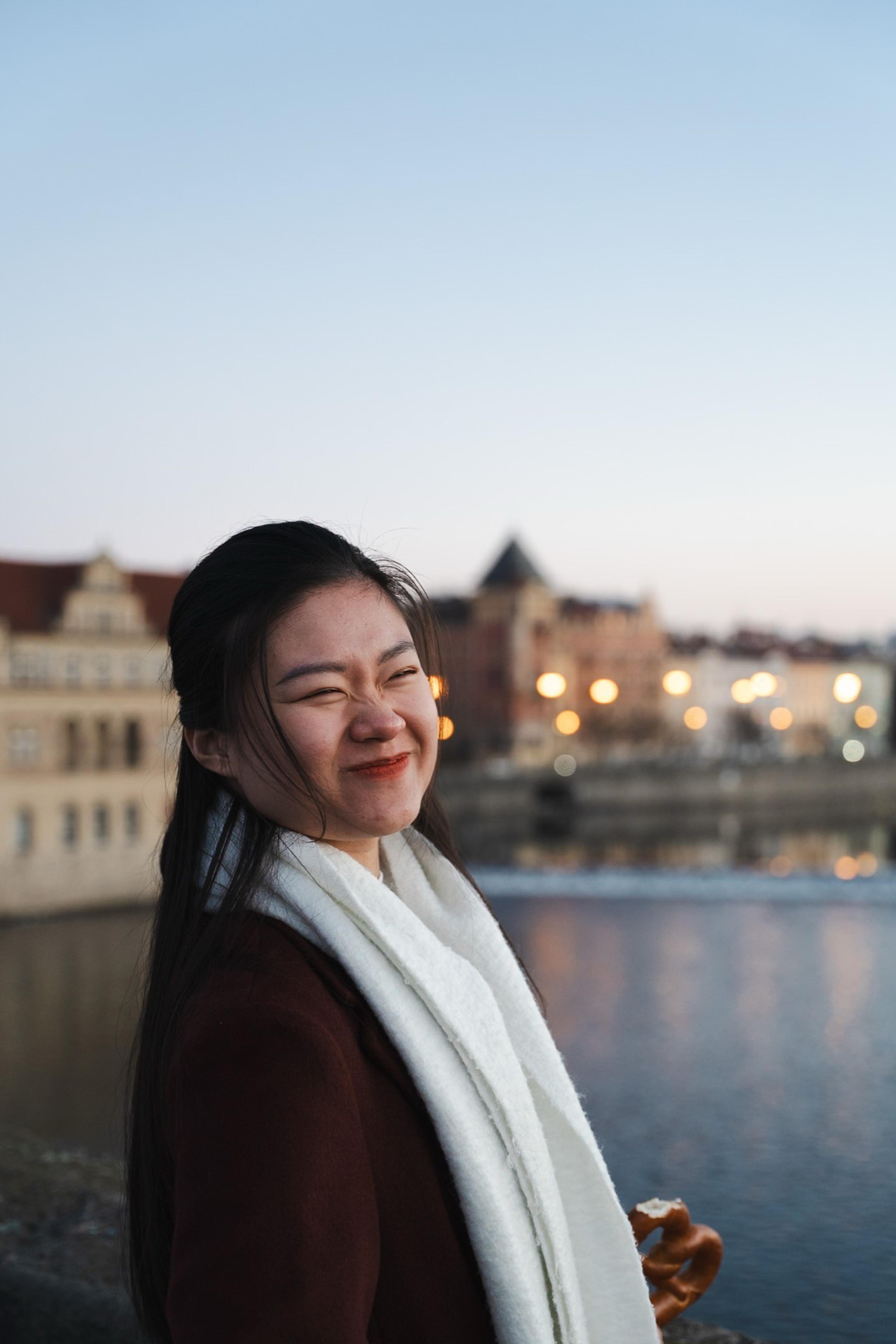 Laughing woman in a white scarf holding a pretzel by a river at dusk, city lights behind