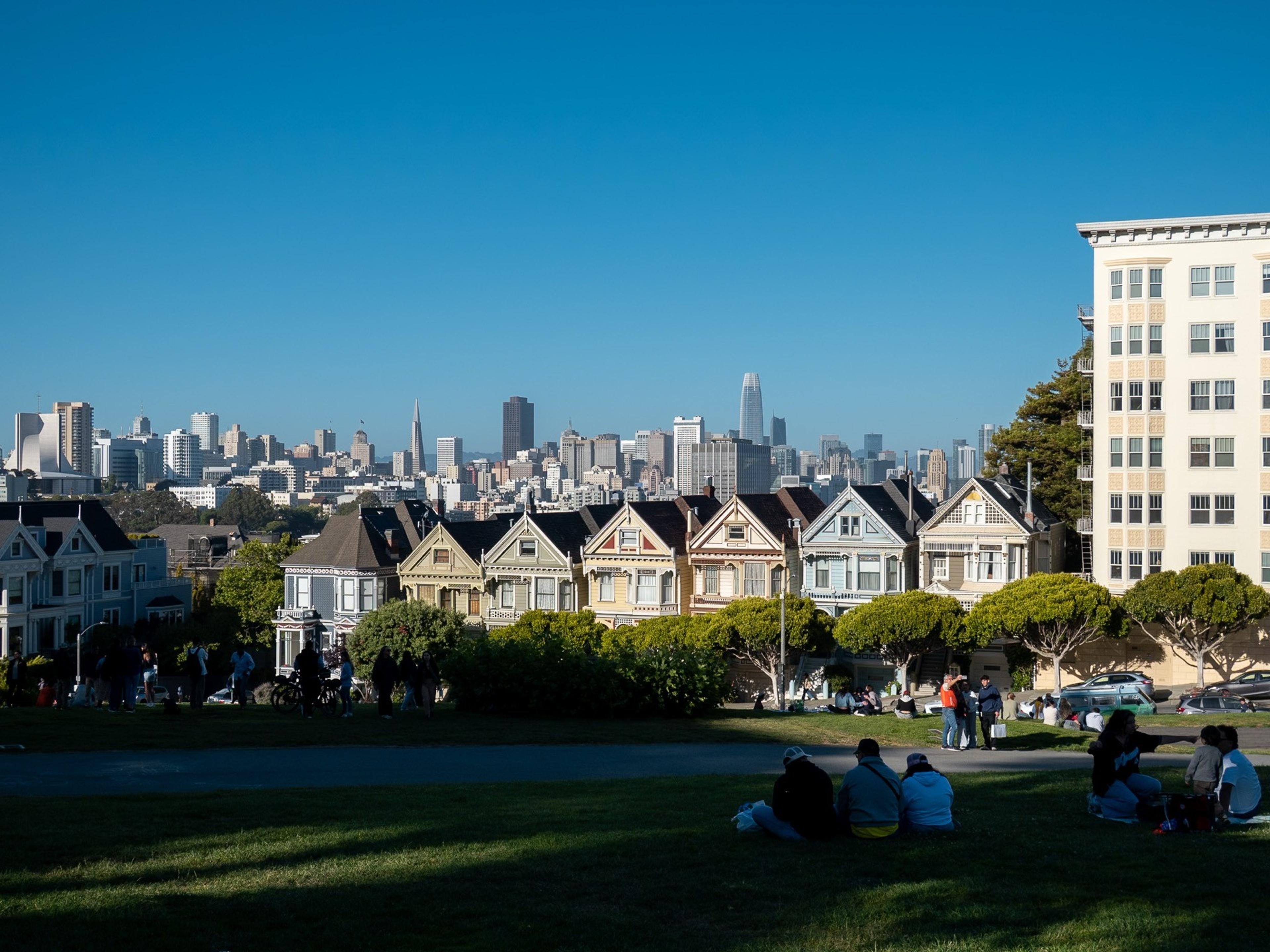 People relaxing on a park lawn before a row of Victorian houses with a downtown skyline behind