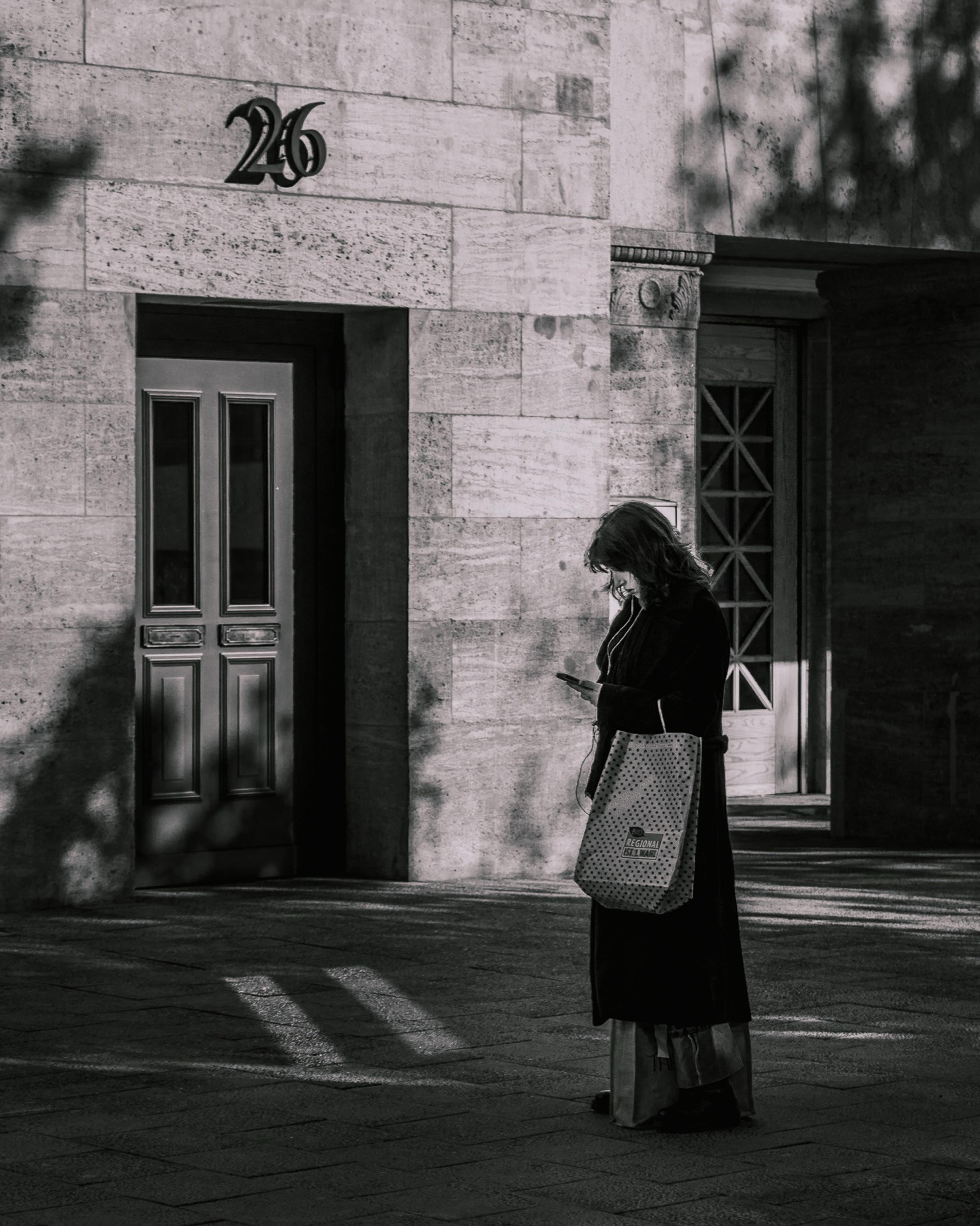 Black and white shot of a woman checking her phone with a tote bag outside a stone building numbered 26