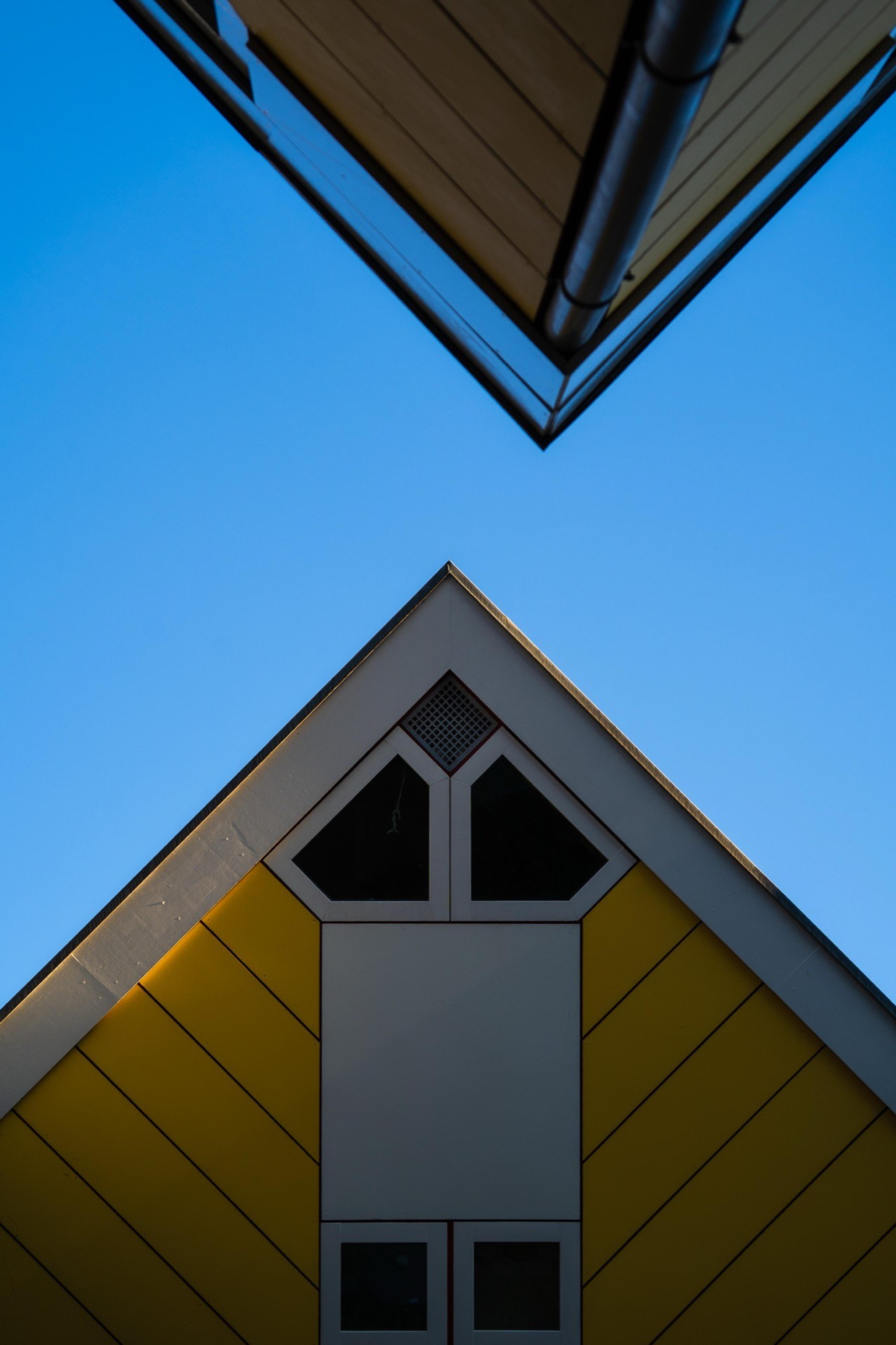 Looking up between two tilted yellow and white cube-shaped houses against a clear blue sky