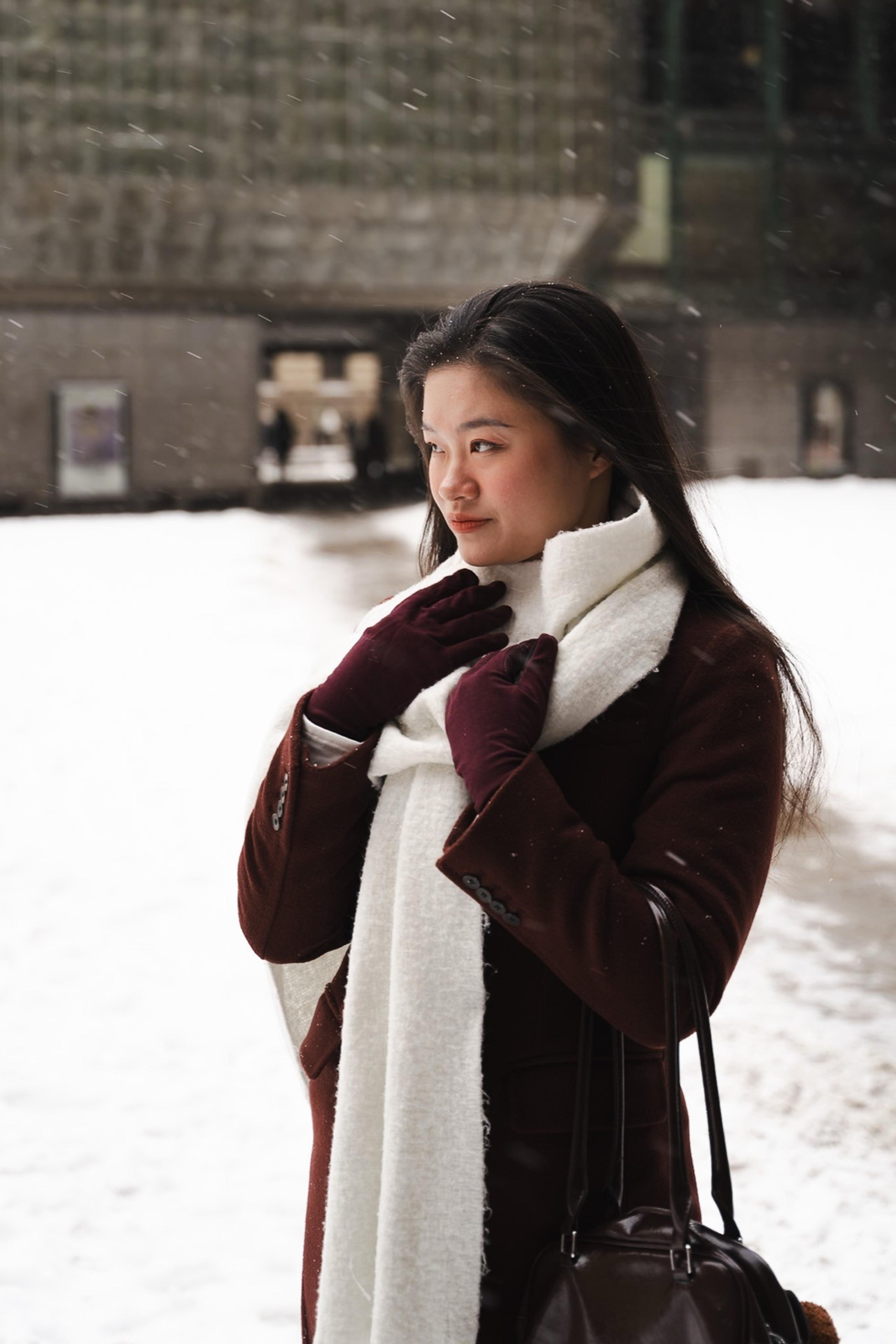 Woman in a burgundy coat, white scarf and gloves standing in falling snow on a city square