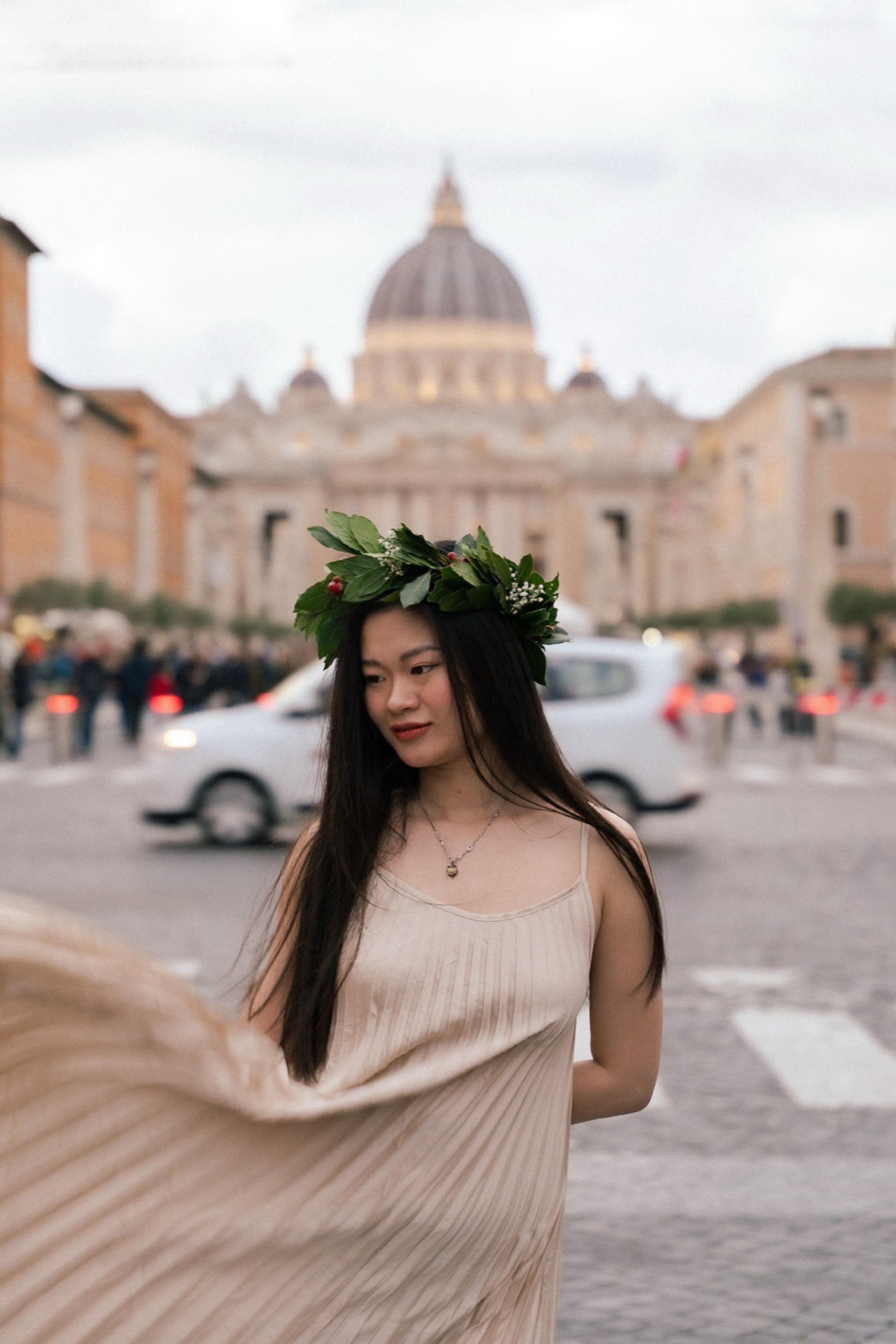 Woman in a leaf crown and pleated dress on a street with a large domed basilica behind her