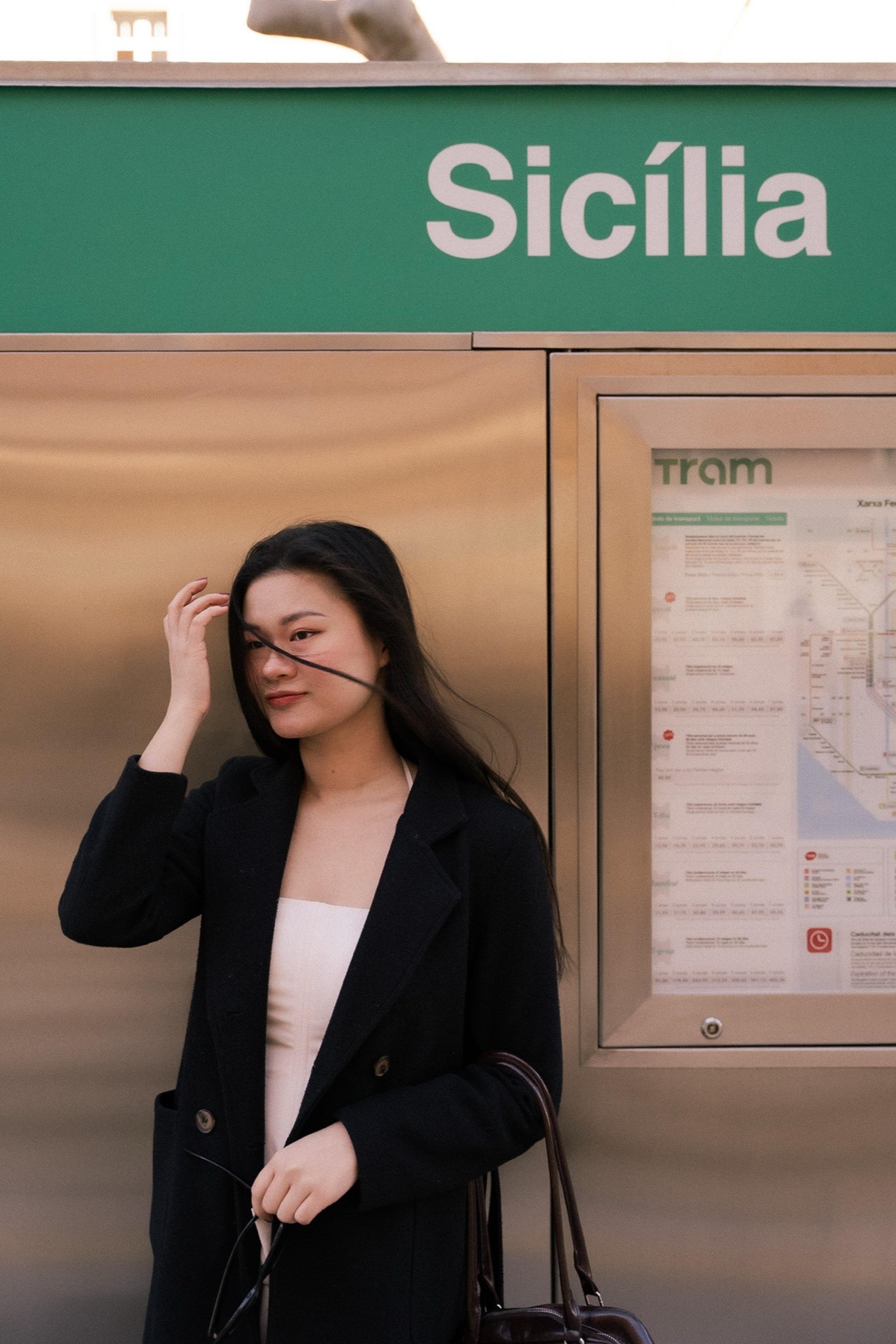 Woman in a black coat posing by a tram stop with a green Sicília sign and a route map