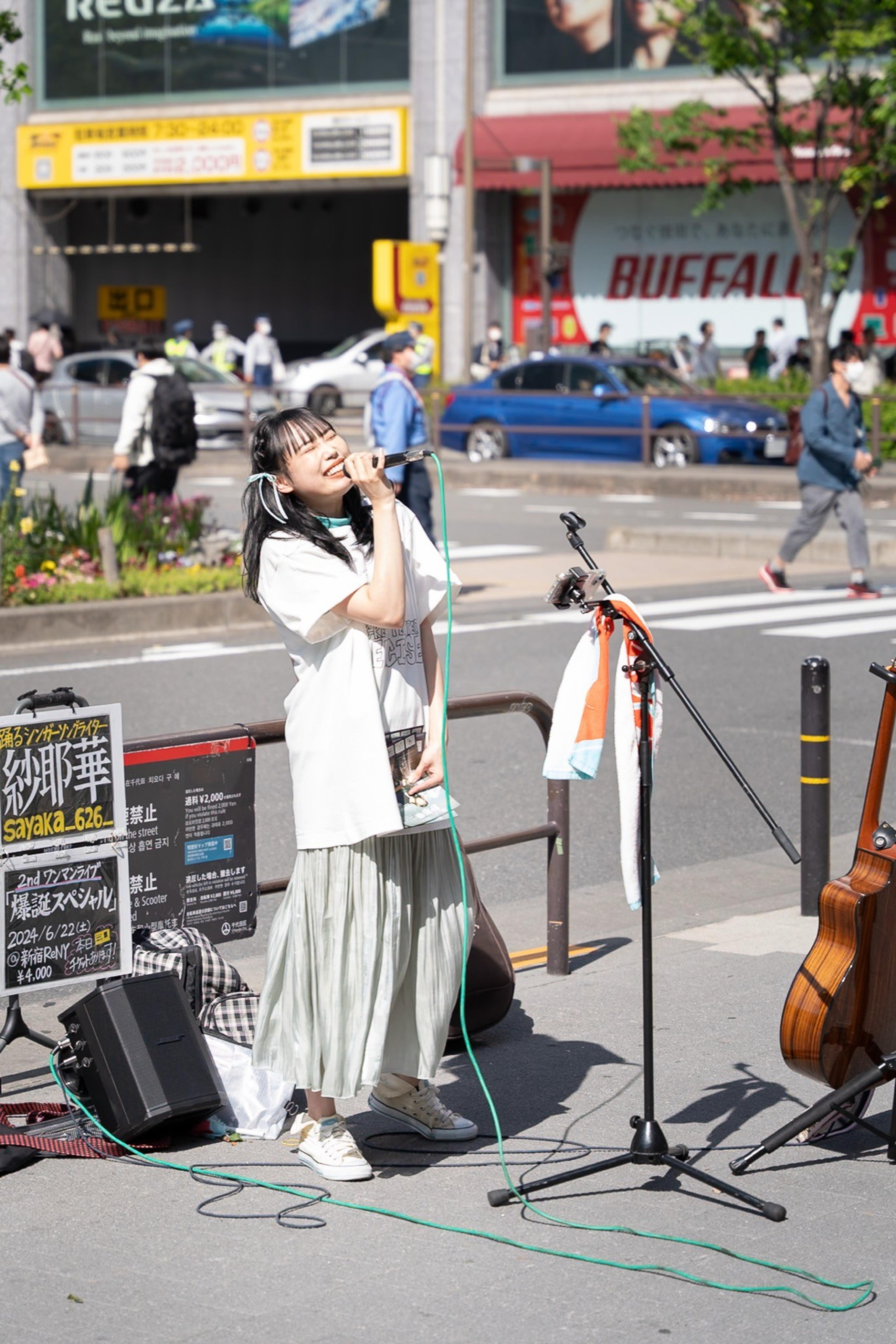Young street singer performing into a microphone on a sunny sidewalk beside a guitar and speaker