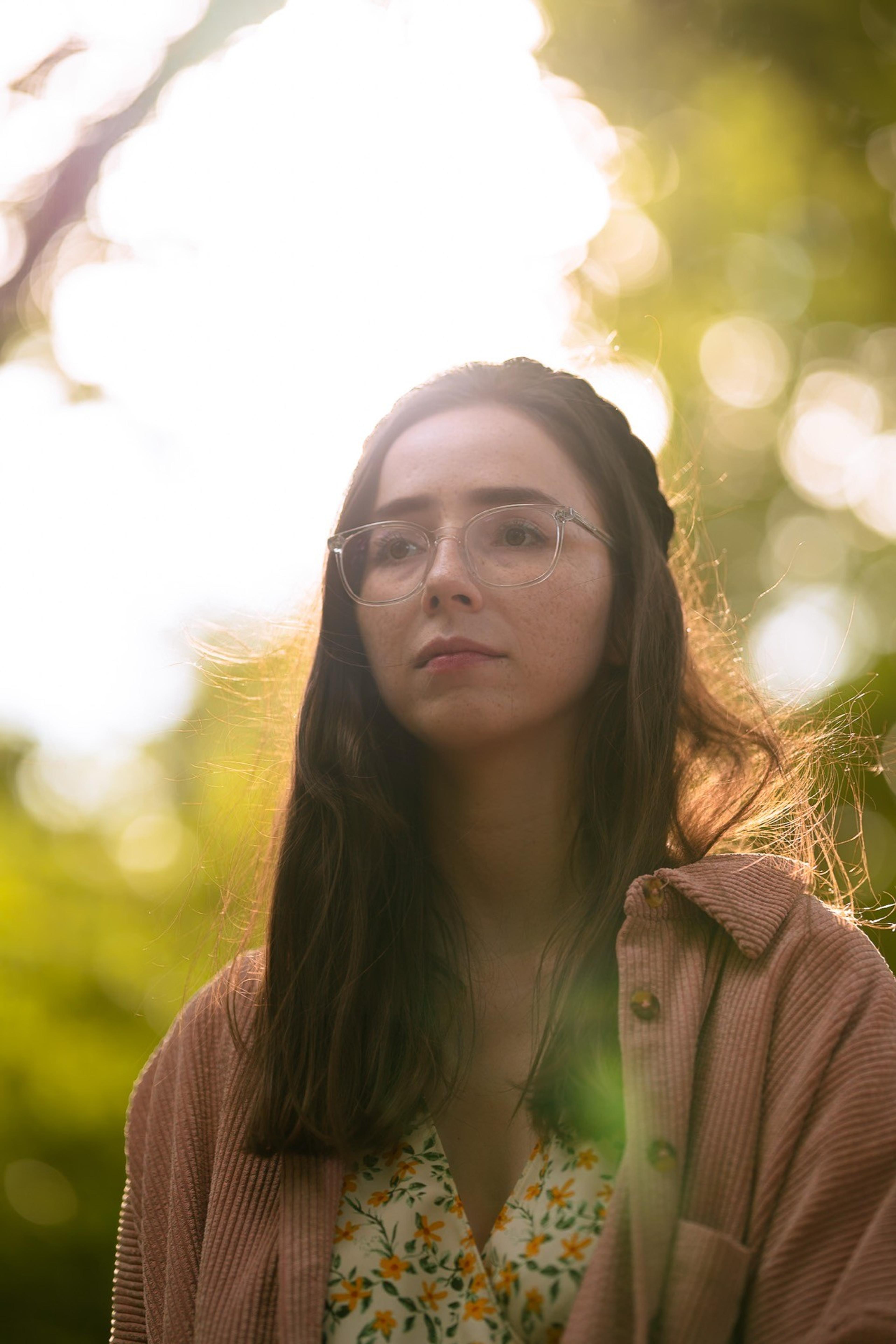 Backlit portrait of a woman with glasses and long hair in a pink jacket among green trees