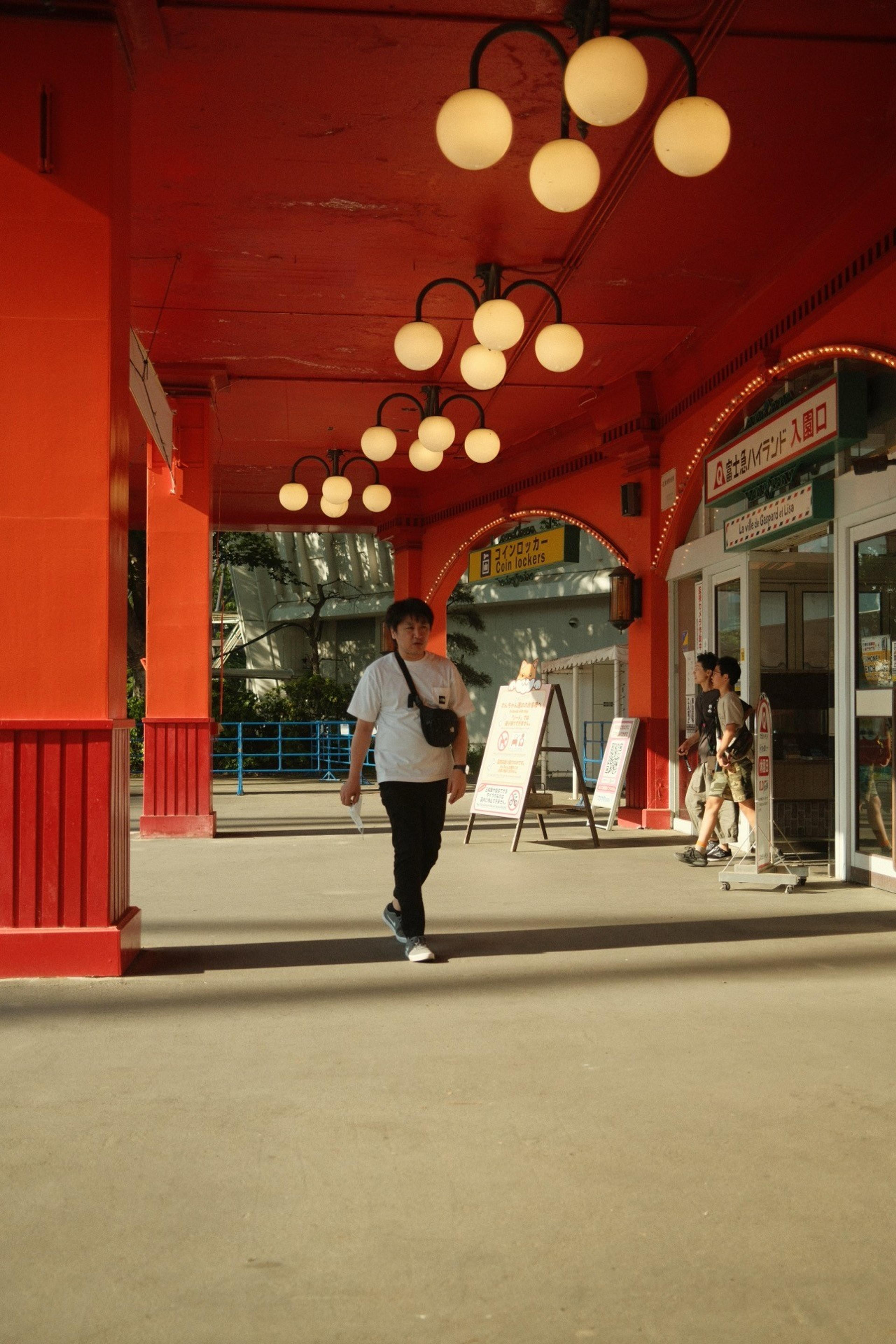 Man walking under a red arcade with globe lamps past an entrance with Japanese signs