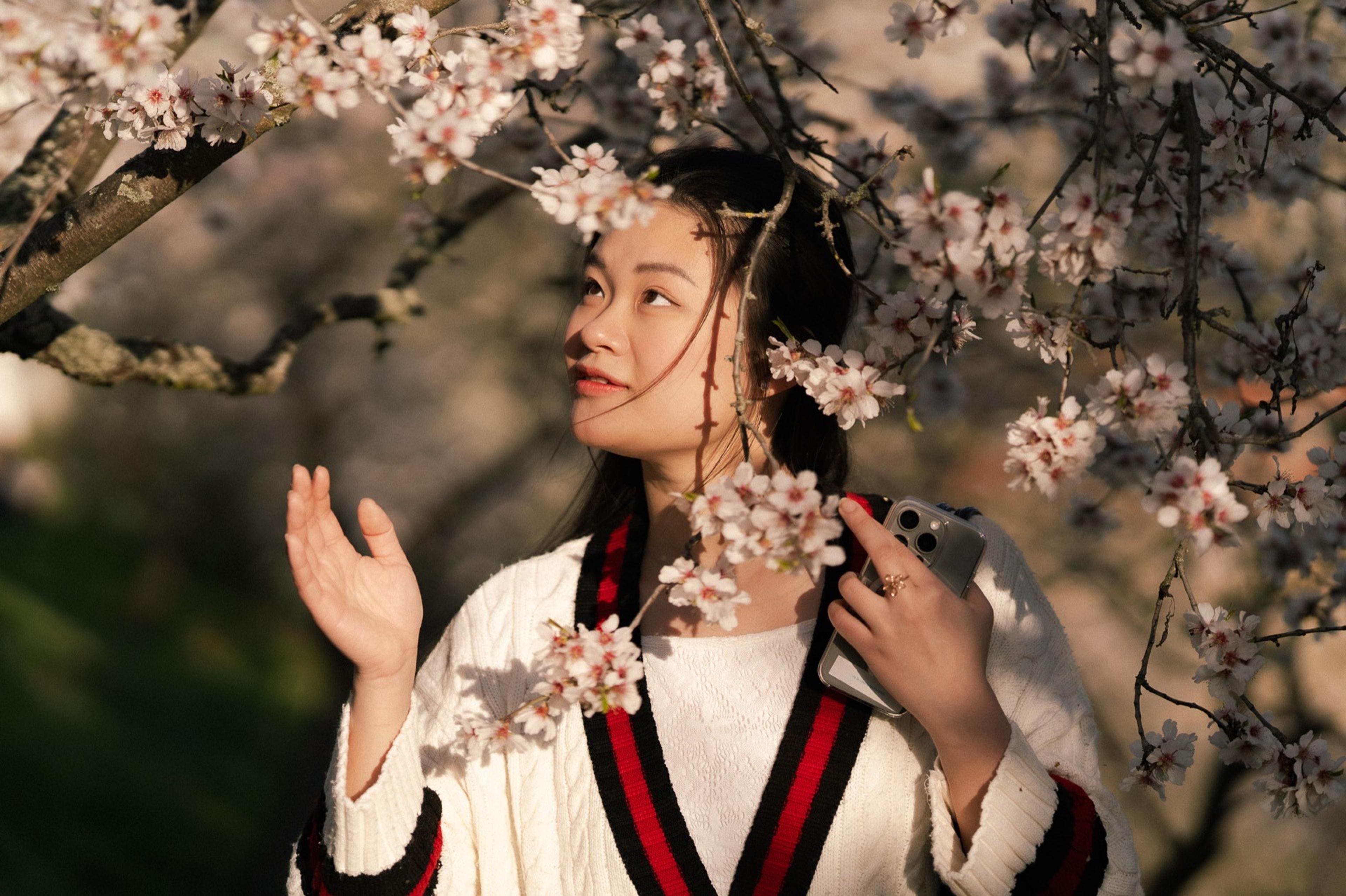 Woman in a white sweater holding a phone, looking up beneath branches of blossoming trees