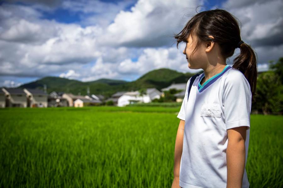 A little girl stares out into green rice paddies. Here are mountains in the distance and large puffy clouds hanging in the sky. This photo was taken in Kyoto, Japan.