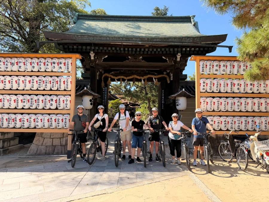 A group of seven cyclists in helmets standing with e-bikes in front of a large wooden shrine gate flanked by walls of inscribed paper lanterns in Kyoto.