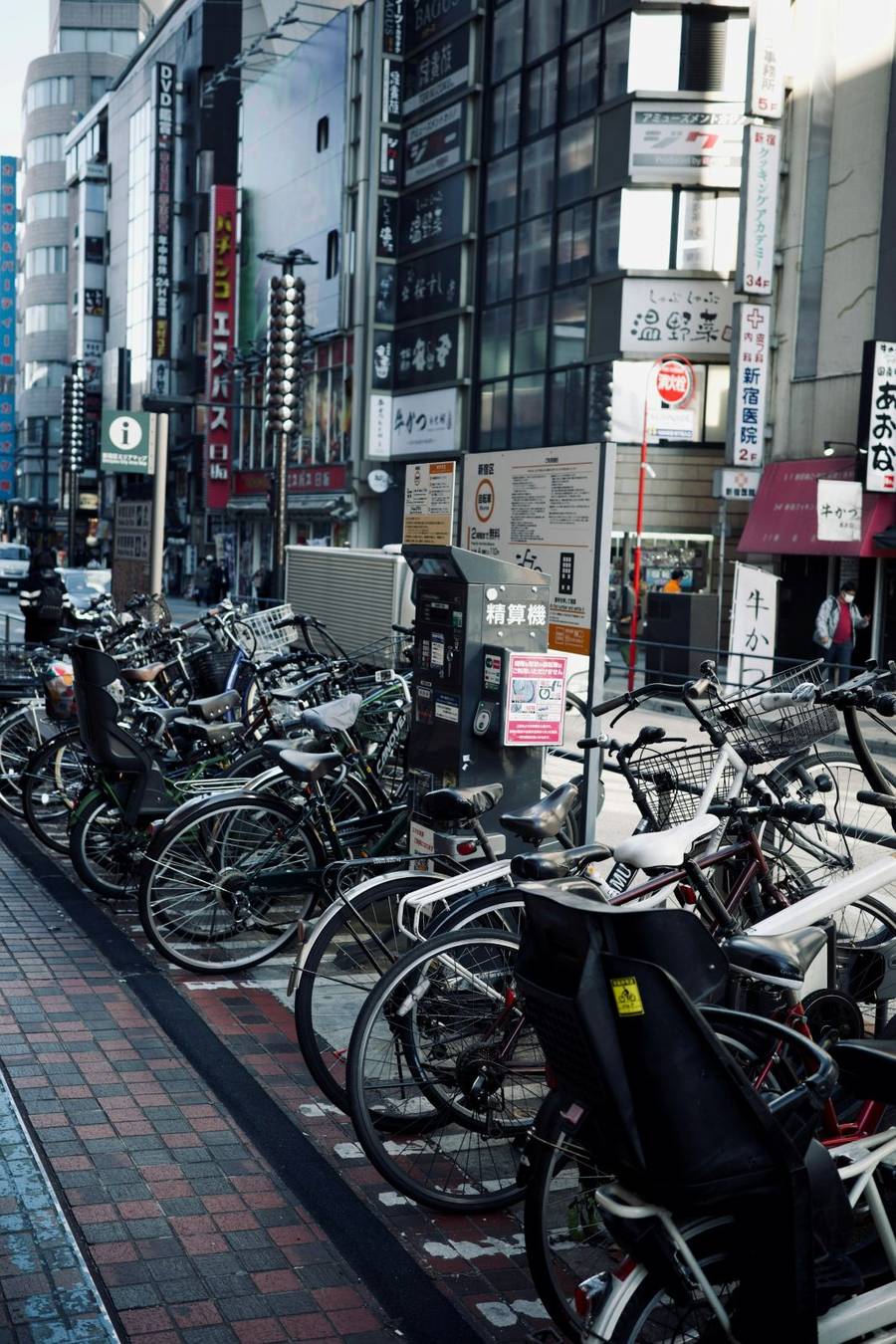 Rows of bicycles parked in a designated paid bicycle parking area on a busy city street in Japan, with a payment machine in the centre and commercial signage in the background.
