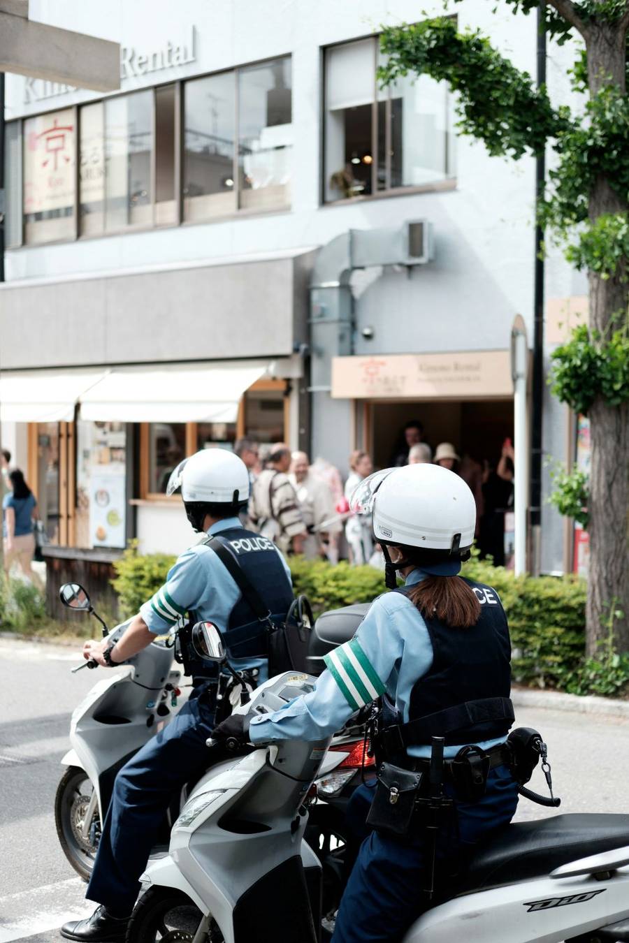 Two Japanese police officers on white scooters parked at a Kyoto intersection, observing a busy street near kimono rental shops.