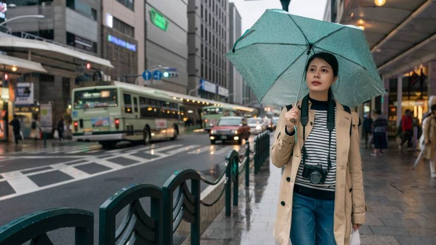 A woman holding a mint-green umbrella walks along a rainy sidewalk on Shijo Street in downtown Kyoto, with a city bus and urban storefronts in the background.