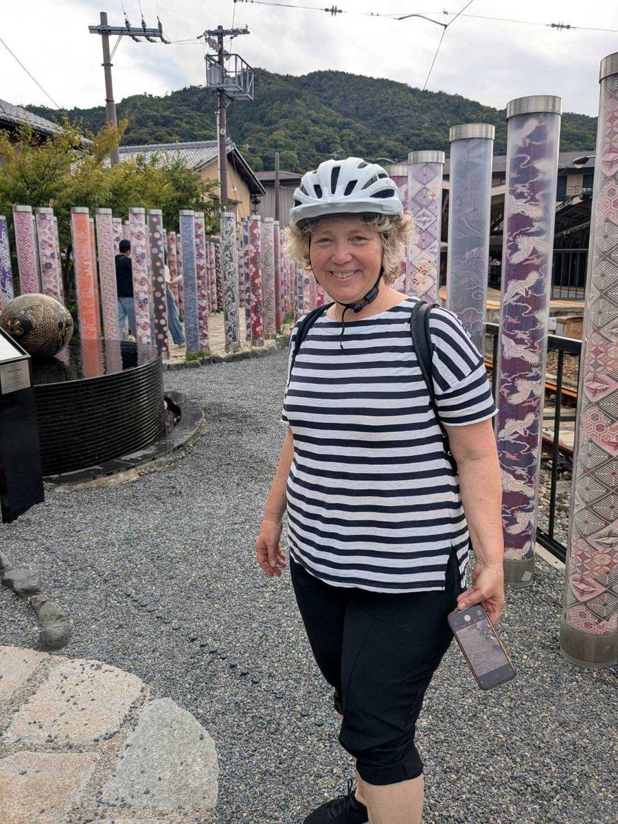 Smiling woman wearing a bike helmet stands among the colorful Kimono Forest pillars at Arashiyama Station with Kyoto's mountains in the background