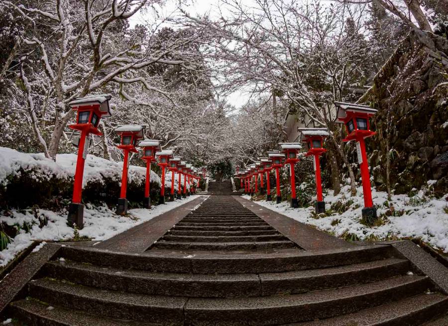 Snow-covered stone steps leading up to Kurama Shrine in Kyoto, lined with traditional red lanterns dusted with fresh snow and bare winter branches overhead.
