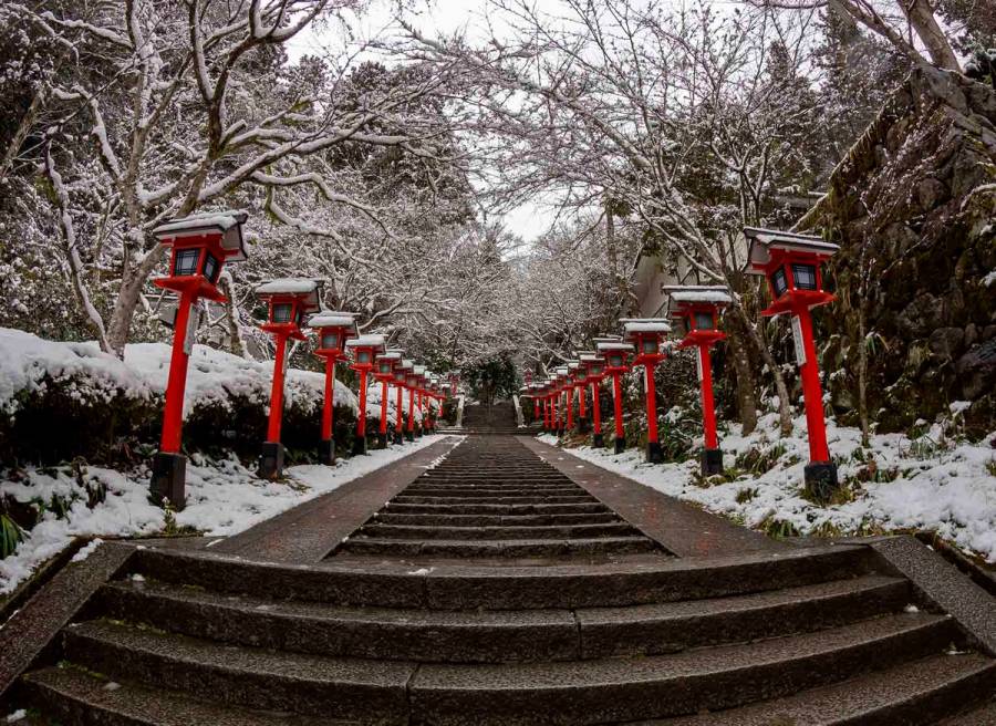 Snow-covered stone steps leading up to Kurama Shrine in Kyoto, lined with traditional red lanterns dusted with fresh snow and bare winter branches overhead.