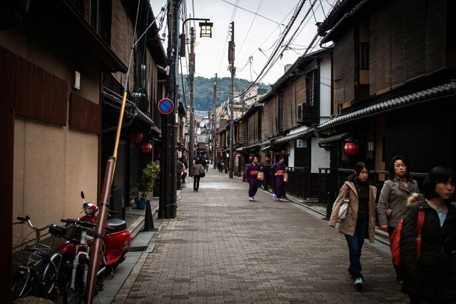 A narrow backstreet in Gion, Kyoto, lined with traditional machiya townhouses and red lanterns, with pedestrians and two women in kimono walking through.