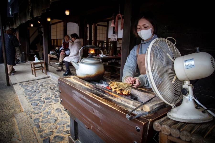 A woman in an apron grills skewered mochi over charcoal at a traditional Japanese tea stall, with customers seated in the background.