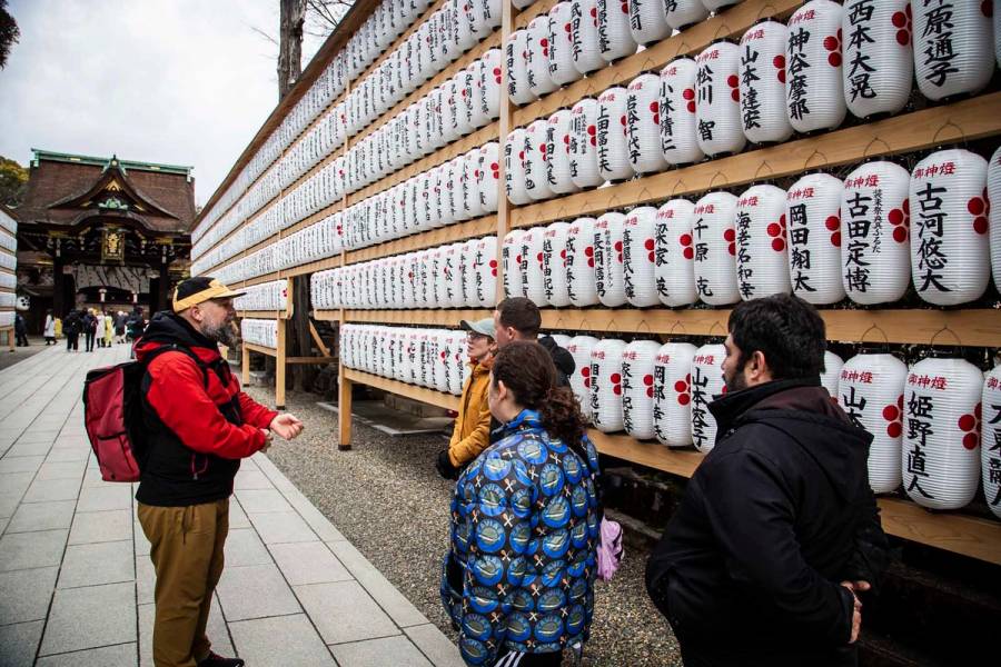 A tour guide in a red jacket gestures while explaining the rows of white paper lanterns inscribed with donor names to a small group of visitors at Kitano Tenmangu shrine on an overcast winter day.