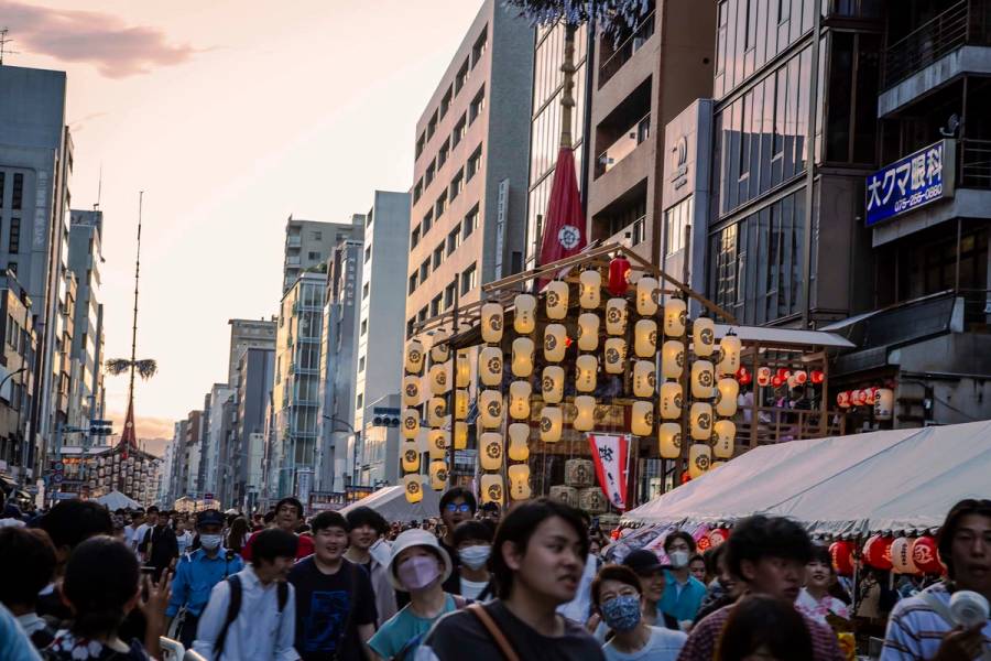 Crowds filling Shijo Street in Kyoto at dusk during the Gion Festival yoi-yama evening, with a large illuminated yamaboko float covered in paper lanterns