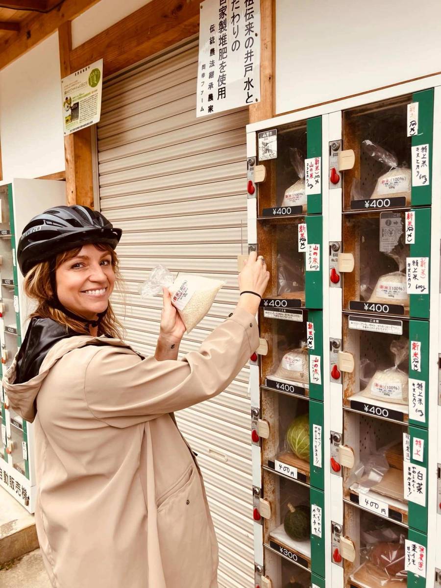 Smiling cyclist wearing a helmet holds a bag of fresh rice purchased from a rural Japanese vending machine selling local produce including rice, vegetables, and cabbage