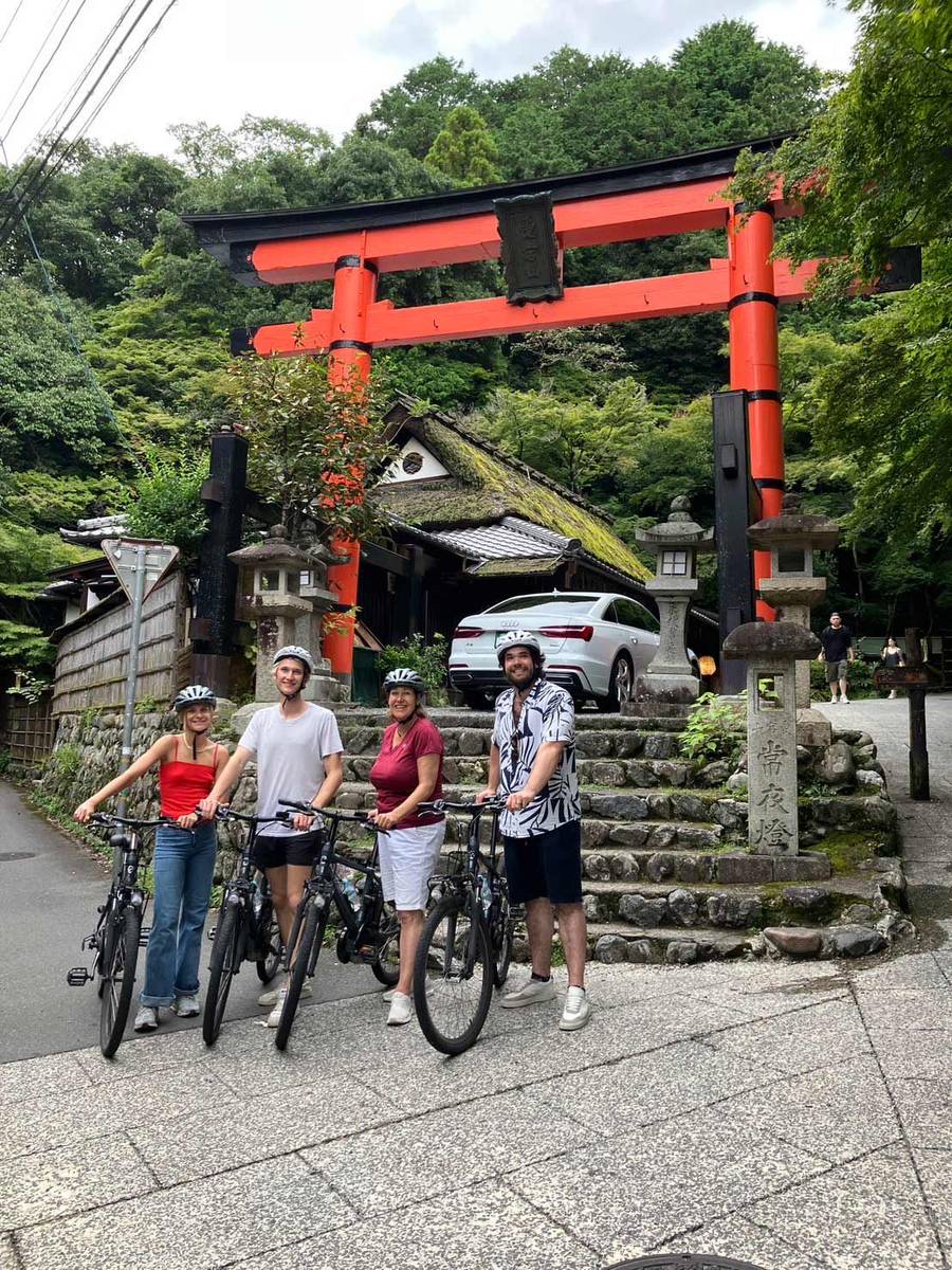 Four happy cyclists with helmets pose with their e-bikes in front of a vermillion torii gate and moss-covered thatched-roof building in Arashiyama, Kyoto