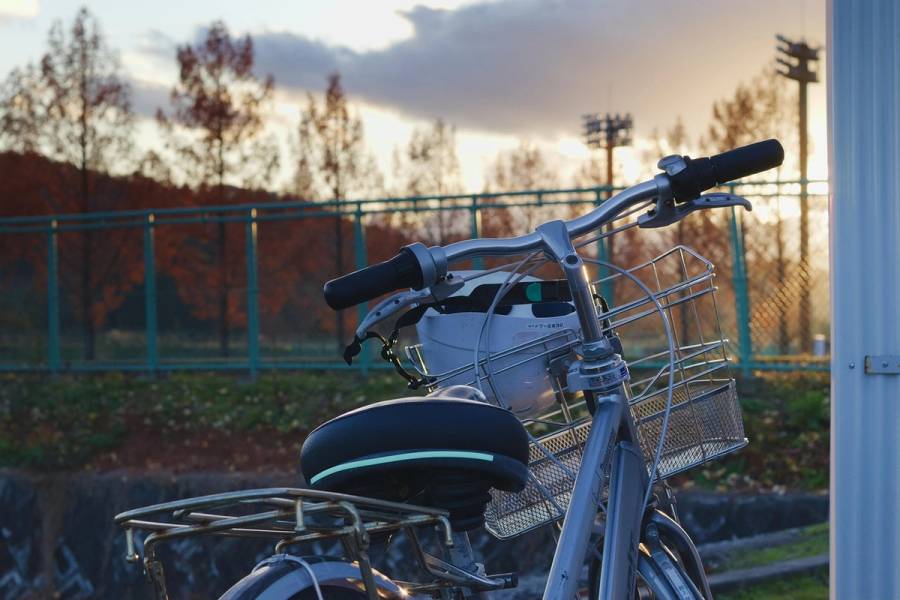 A standard Japanese city bicycle with a front basket parked in front of a fence at sunset, with autumn trees in the background.