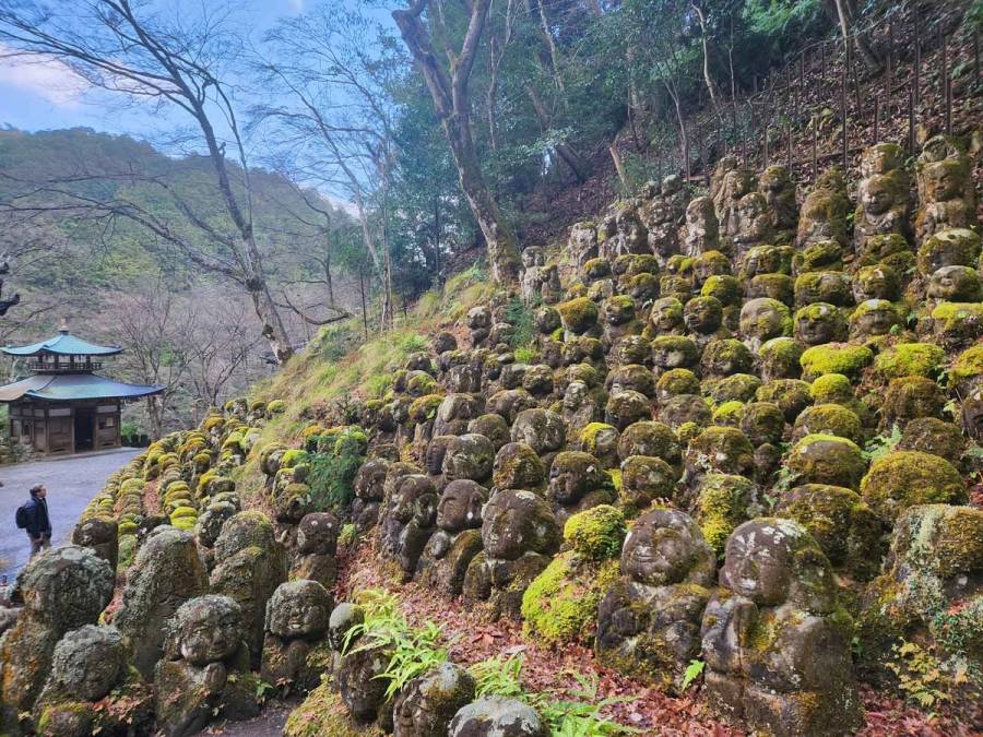 Hundreds of moss-covered stone rakan statues with unique facial expressions line the hillside at Otagi Nenbutsu-ji Temple in Arashiyama, Kyoto, with a traditional pavilion in the background.