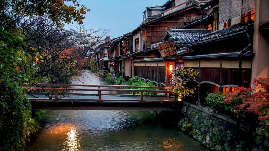 A wooden footbridge over the Shirakawa canal in Kyoto at dusk, lined with traditional machiya buildings and autumn leaves.