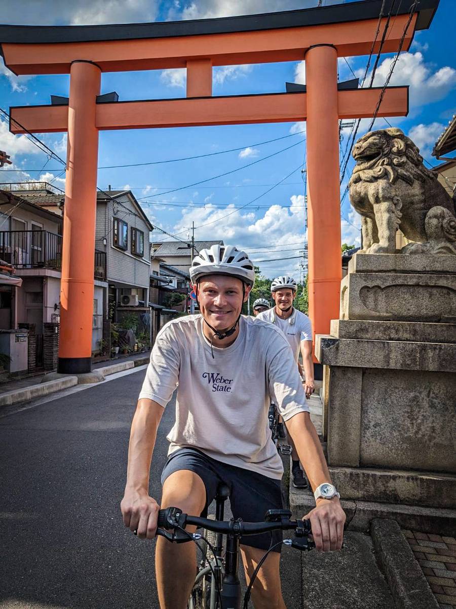 Two cyclists wearing helmets ride through the vermillion torii gate at Imamiya Shrine in Kyoto, passing a stone komainu guardian statue on a quiet neighborhood street.