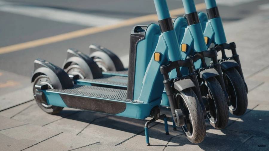 Three blue electric scooters parked on a pavement beside a road, showing the deck, front suspension, and tyres typical of share-ride e-scooter services.