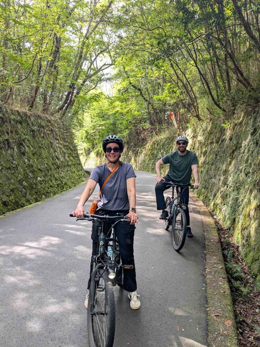 Two smiling cyclists ride e-bikes along a shaded path cut through moss-covered stone walls in the hills of Arashiyama, Kyoto, with a green forest canopy overhead.