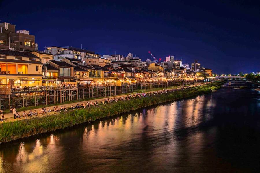 Kamo River at night with illuminated Pontocho yuka restaurant platforms reflected in the water and crowds sitting on the riverbank, Kyoto