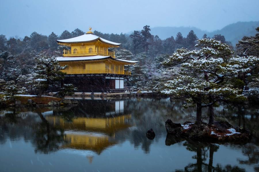 The Golden Pavilion of Kinkaku-ji dusted in snow, reflected in the still pond in front of it as snow falls.