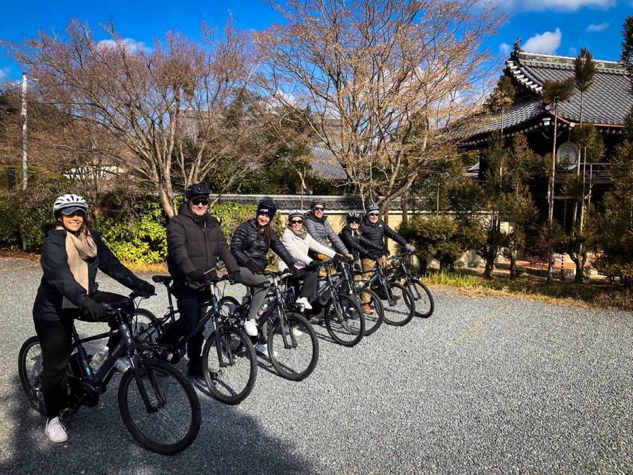 Six tourists wearing helmets seated on e-bikes in a row outside a traditional Japanese temple building on a clear winter day, on a NORU guided bike tour in Kyoto.