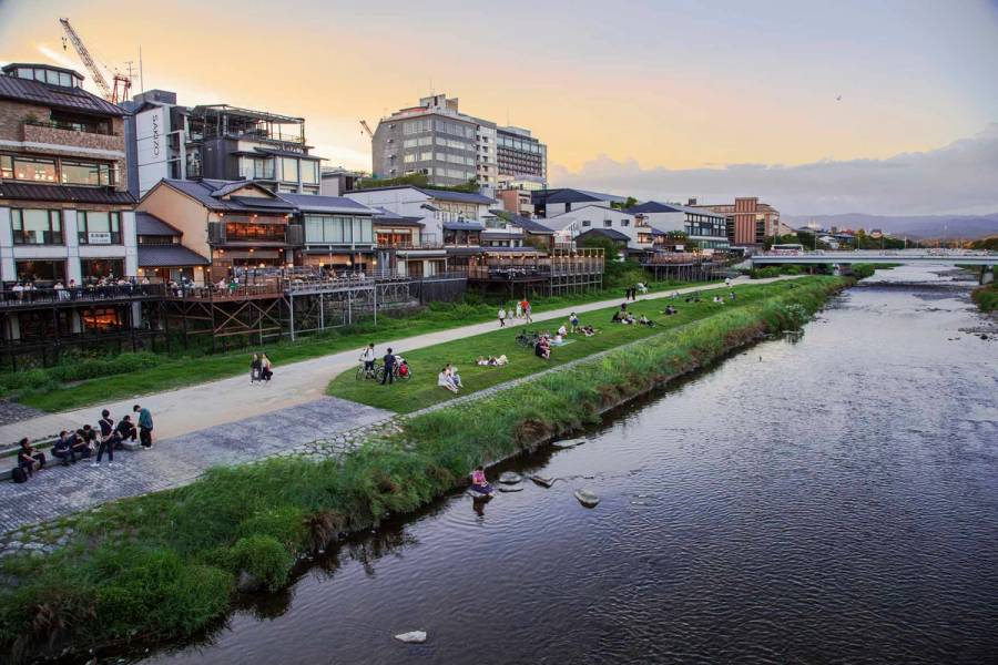 Evening view of Kyoto's Kamo River near Sanjo Bridge, with locals relaxing on grassy riverbanks and traditional kawayuka dining platforms extending from riverside restaurants at sunset.
