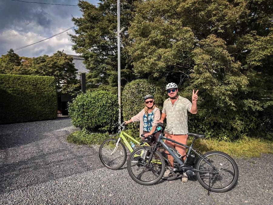 A smiling couple poses with their e-bikes on a quiet residential street in Kyoto, wearing helmets and sunglasses with traditional Japanese hedges and greenery behind them.