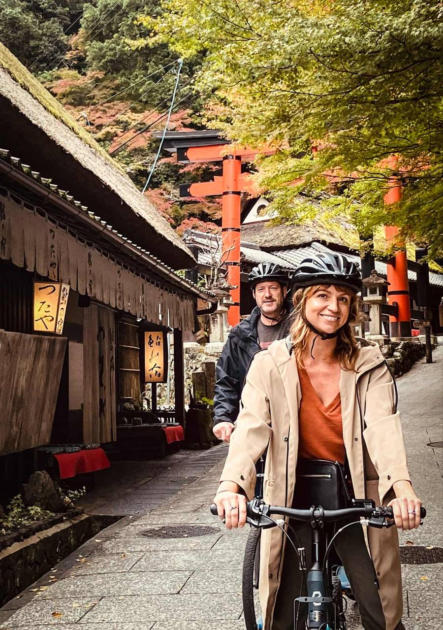 Two cyclists wearing helmets ride along a quiet stone path in Arashiyama, passing traditional thatched-roof buildings and vermillion torii gates surrounded by autumn foliage in shades of green and orange.